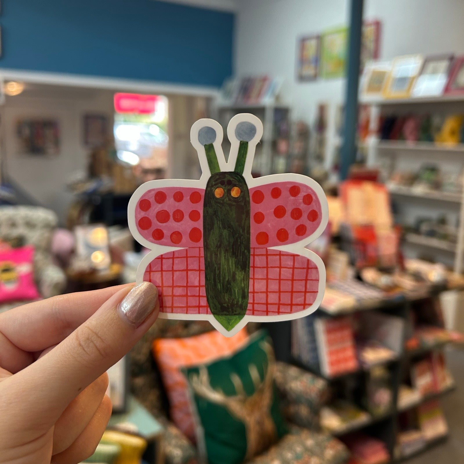 A hand holds a sticker shaped like a butterfly with red polka-dot wings and a green striped body, set against a blurred shop background.