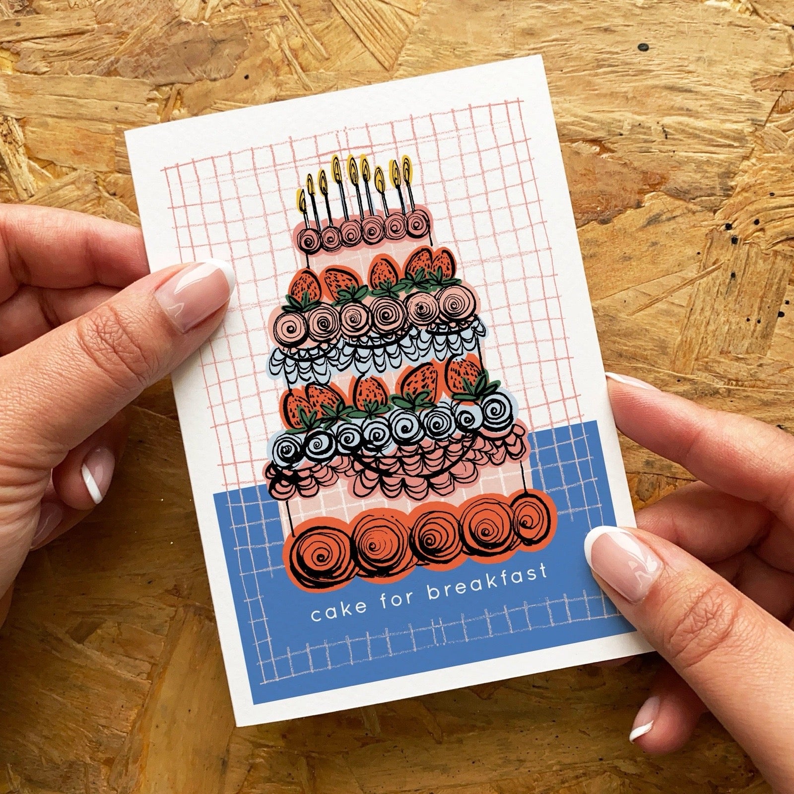 Card with a colourful cake illustration held by hands on a wooden surface.