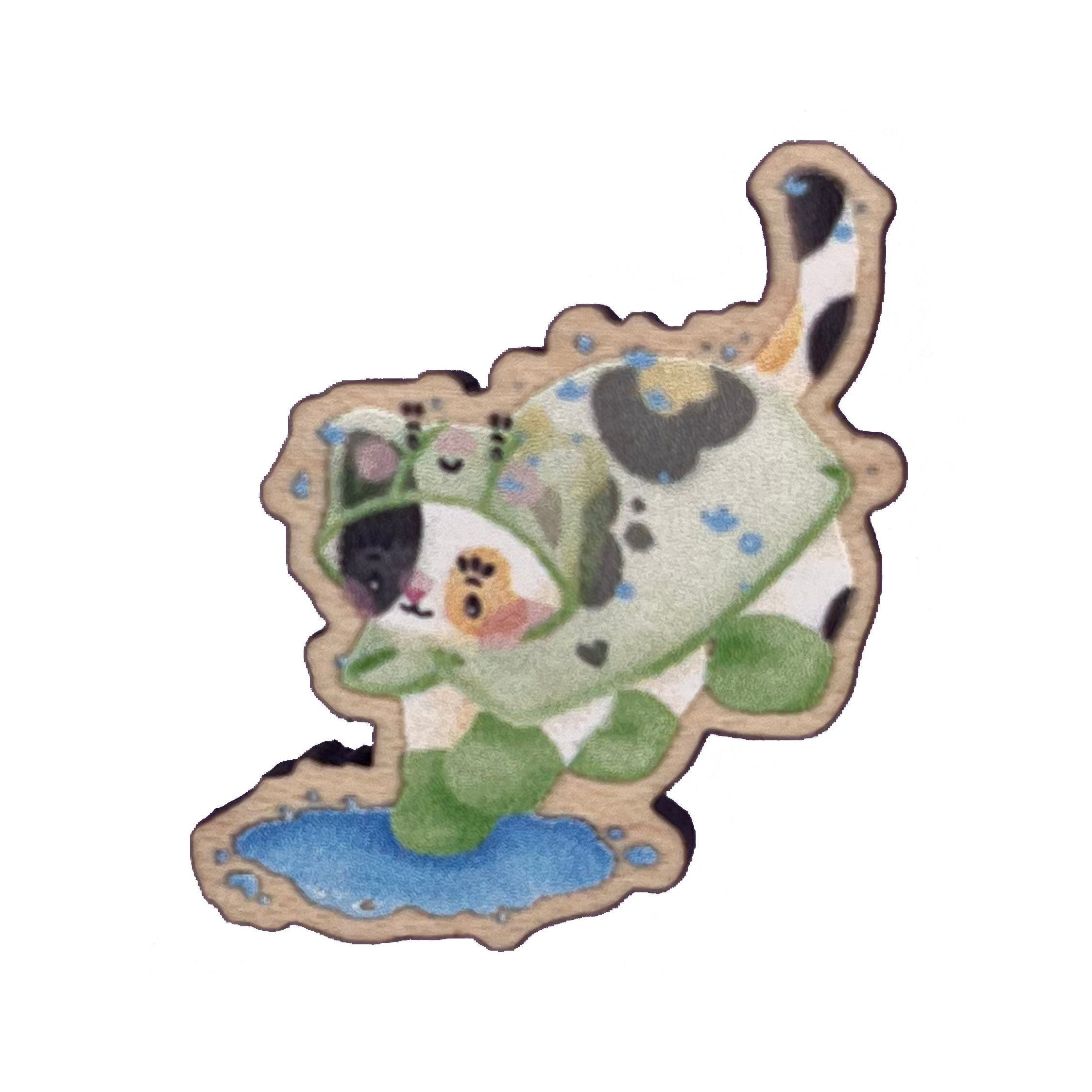 Wooden pin badge of a calico cat wearing a frog raincoat and green rainboots, jumping into a puddle. This badge is printed onto 4mm thick responsibly sourced UK maplewood. The badge has a pink, heart-shaped, rubber backing.