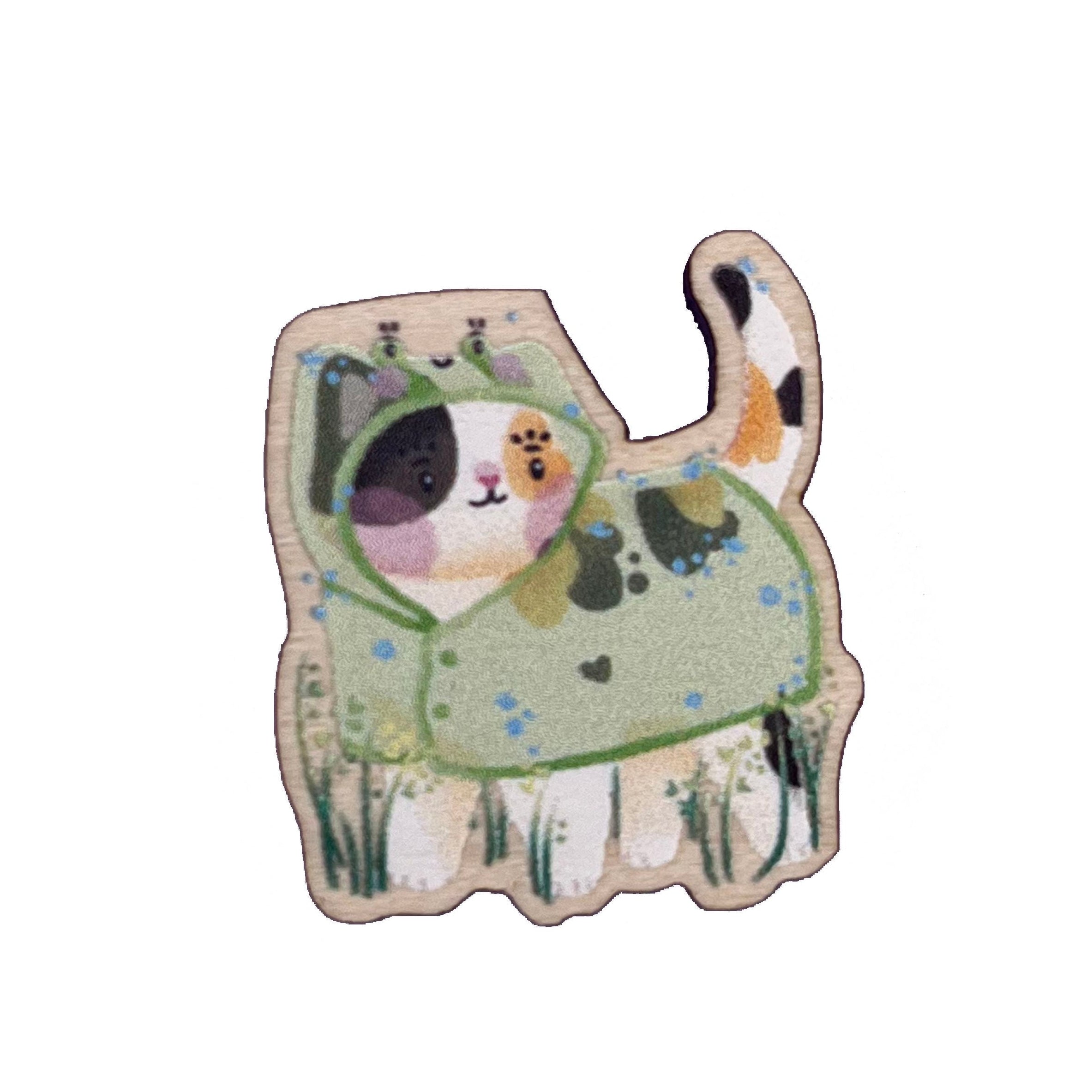 Wooden pin badge of a calico cat wearing a frog raincoat. This badge is printed onto 4mm thick responsibly sourced UK maplewood. The badge has a pink, heart-shaped, rubber backing.