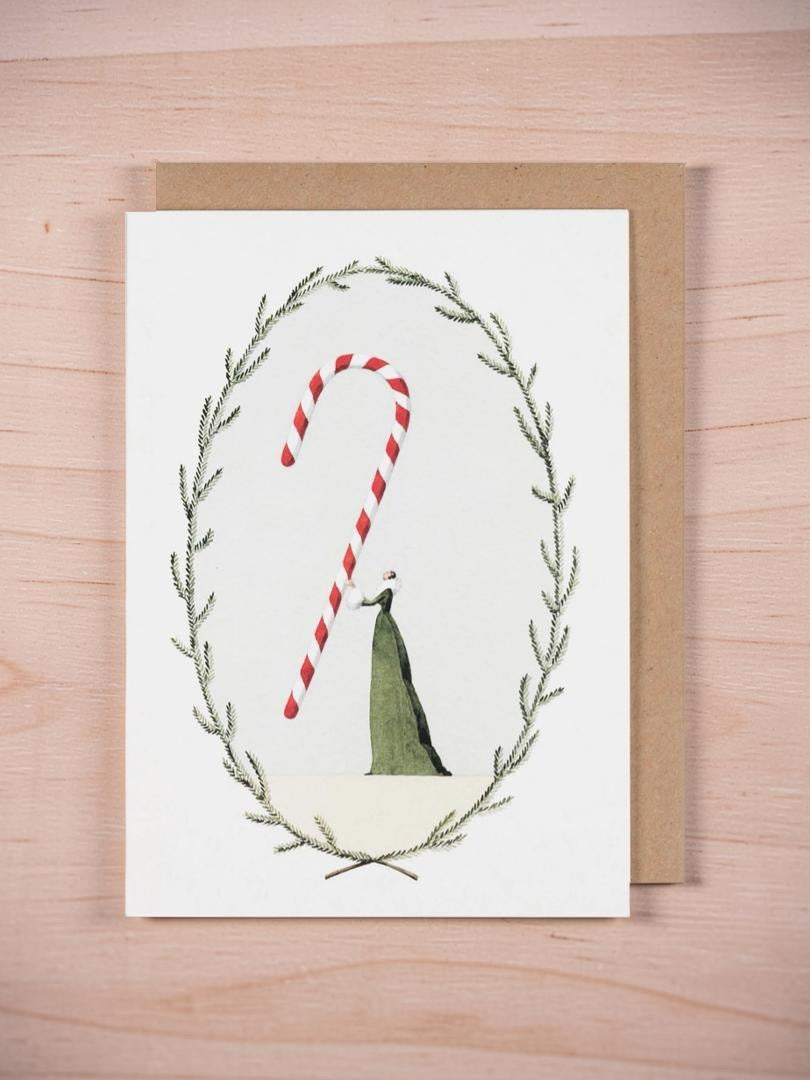 Christmas card featuring a lady in a beautiful green dress holding a giant red and white striped candy cane, framed by green foliage, on wooden background.