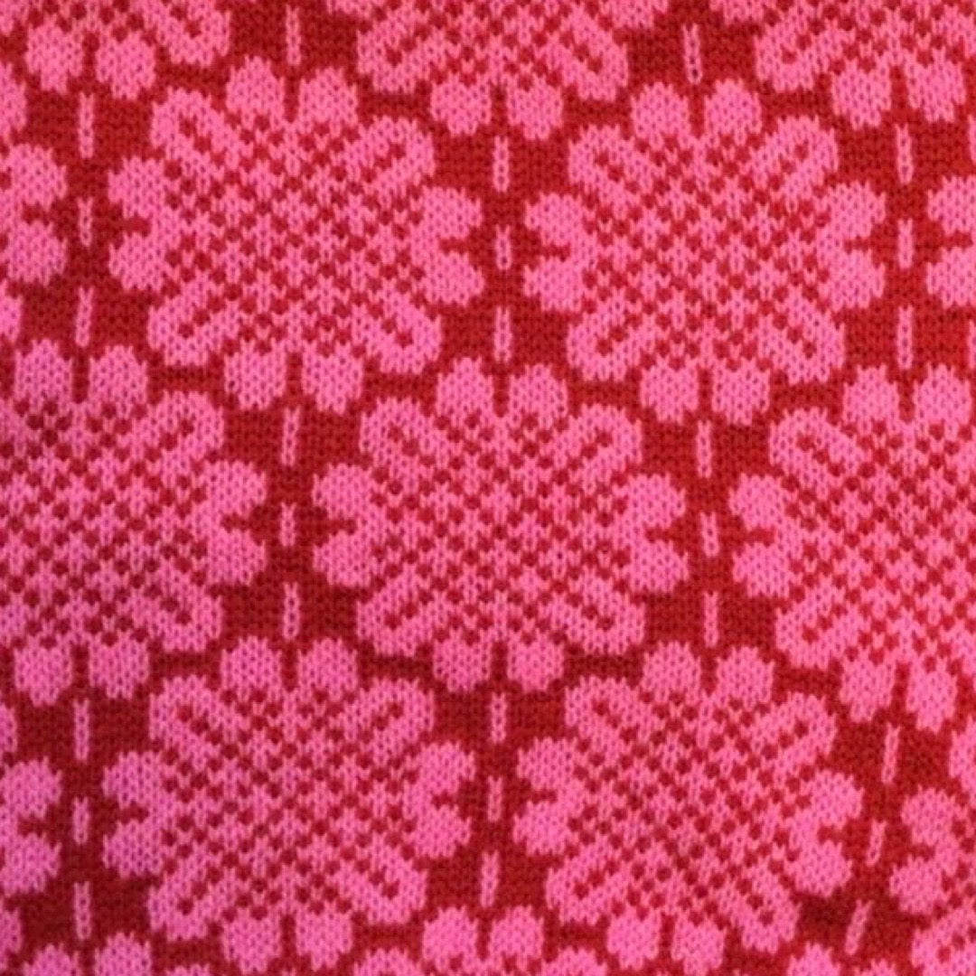 A soft and cosy 100% lambswool knitted snood in a pink daisy design against a red background.