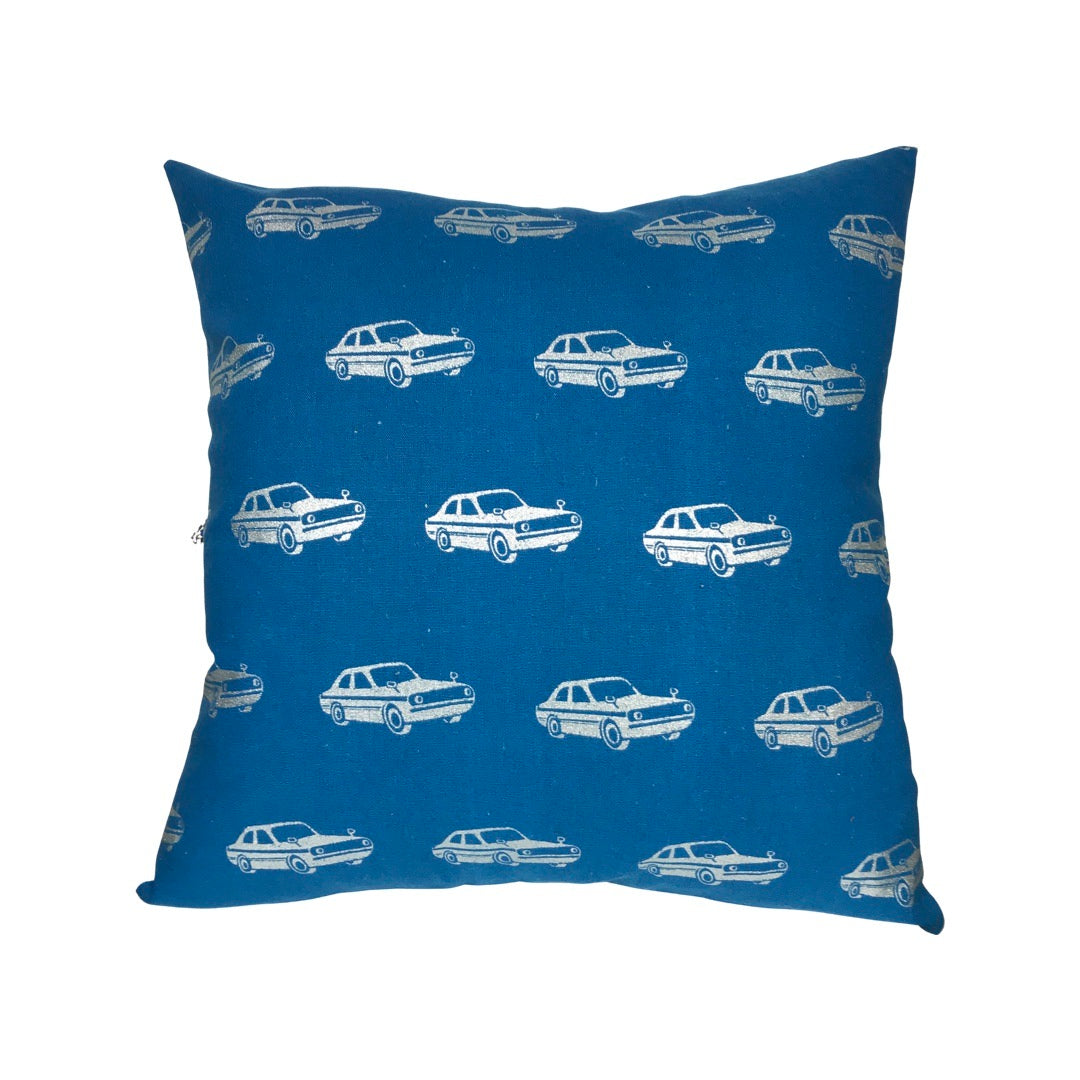 A square cushion cover with a blue background featuring metallic silver retro cars pattern, and a grey cotton back with a blue zip.