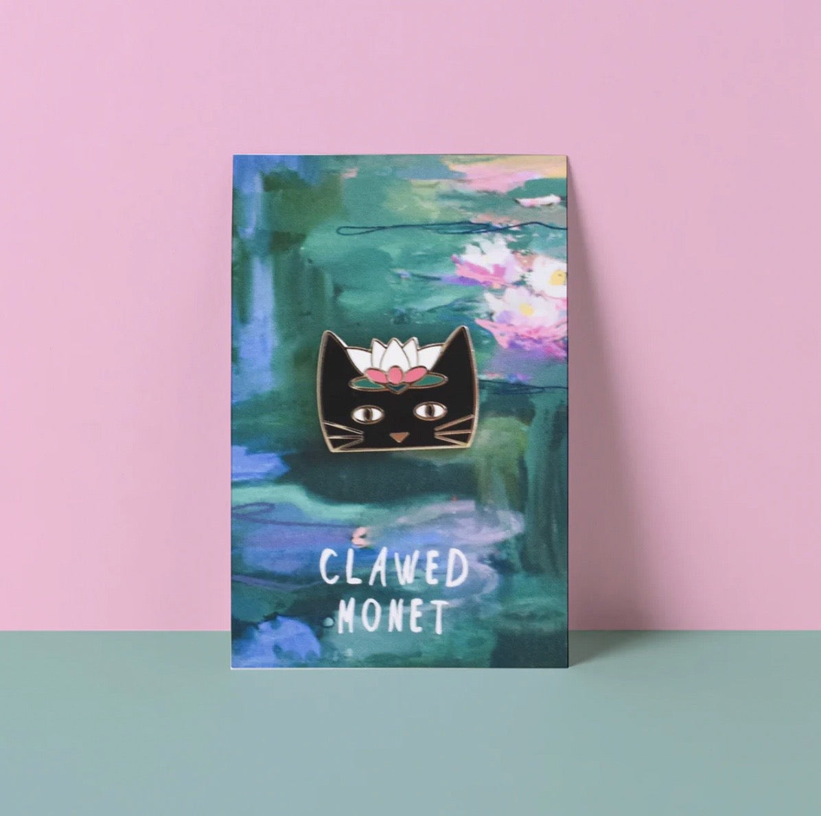 Enamel pin with a black cat design on a card with water lily painting, against a pink and green background.