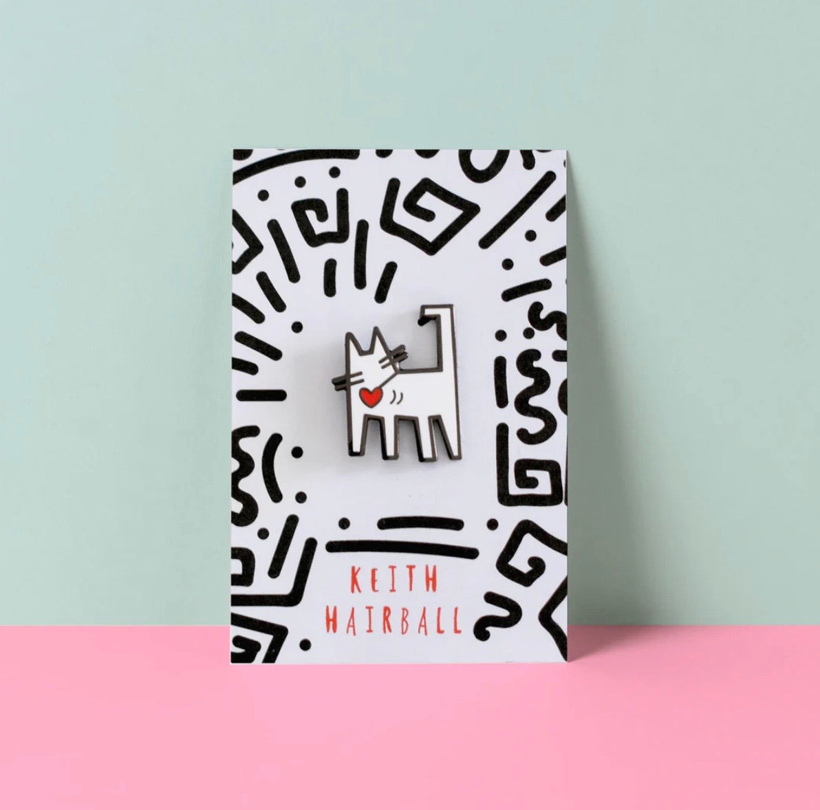 Pin with a cat design on a patterned card against a light blue and pink background.