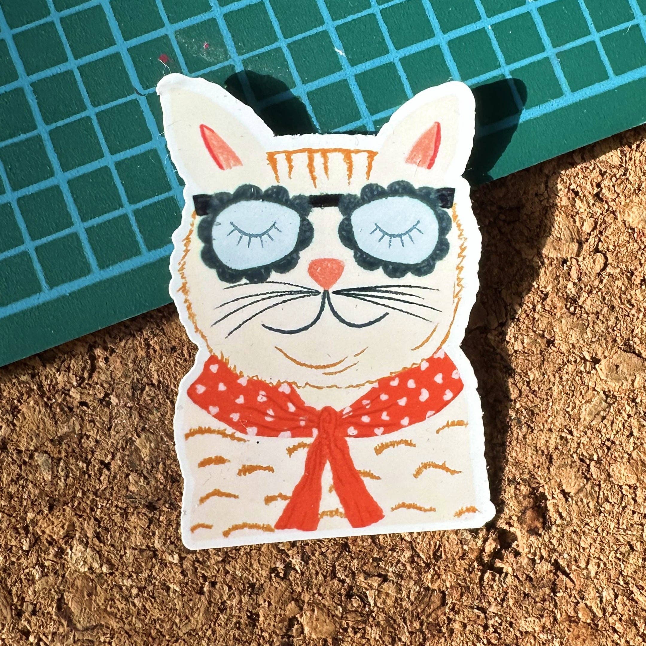 Cat-shaped sticker with sunglasses and a red bow tie on a cork surface with a green mat.