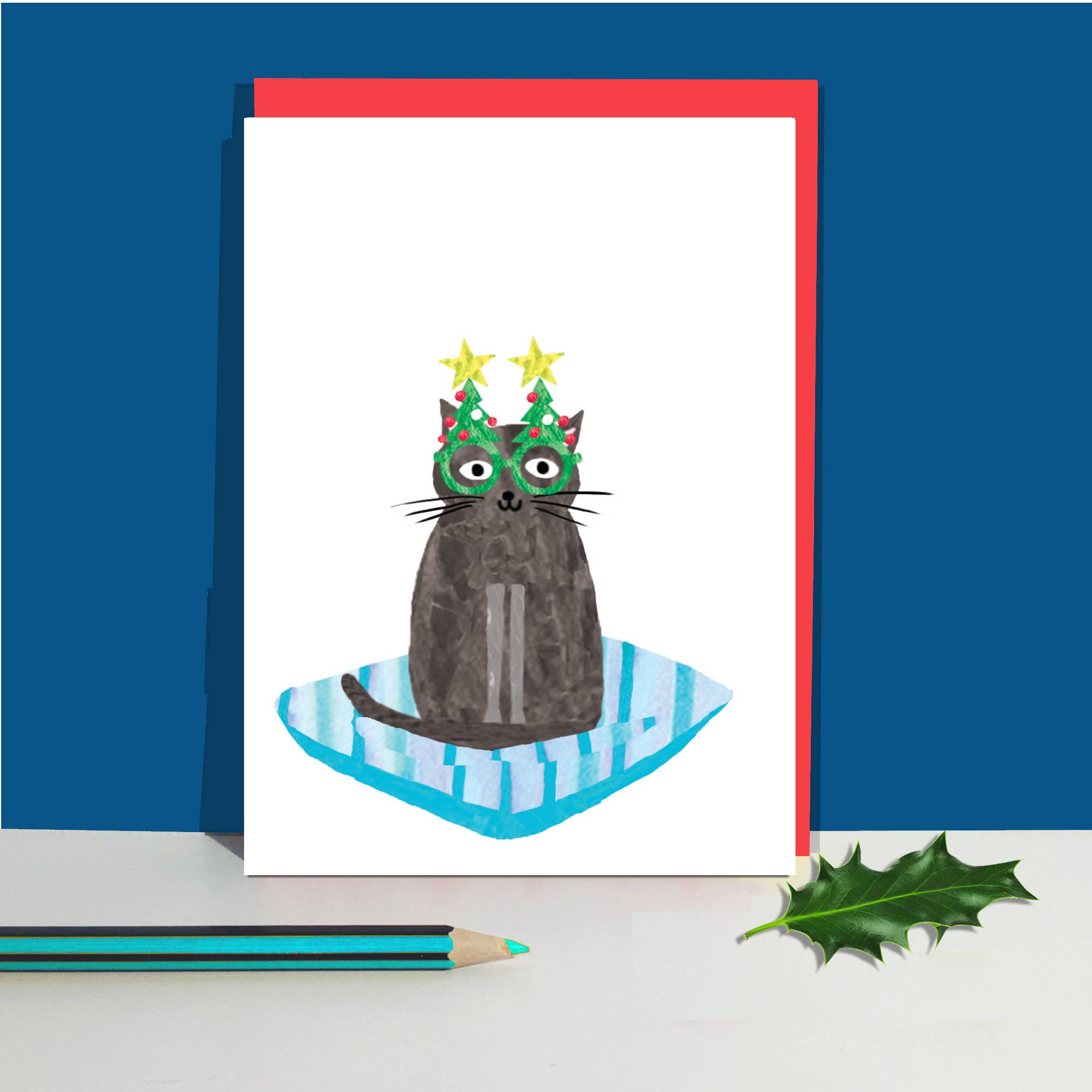 A white greeting card with a whimsical illustration of a dark grey cat wearing a green Christmas hat with yellow stars, sitting on a blue striped cushion. The card is displayed standing upright on a white surface next to a green holly leaf and a teal pencil, with a red envelope behind it against a solid blue background.