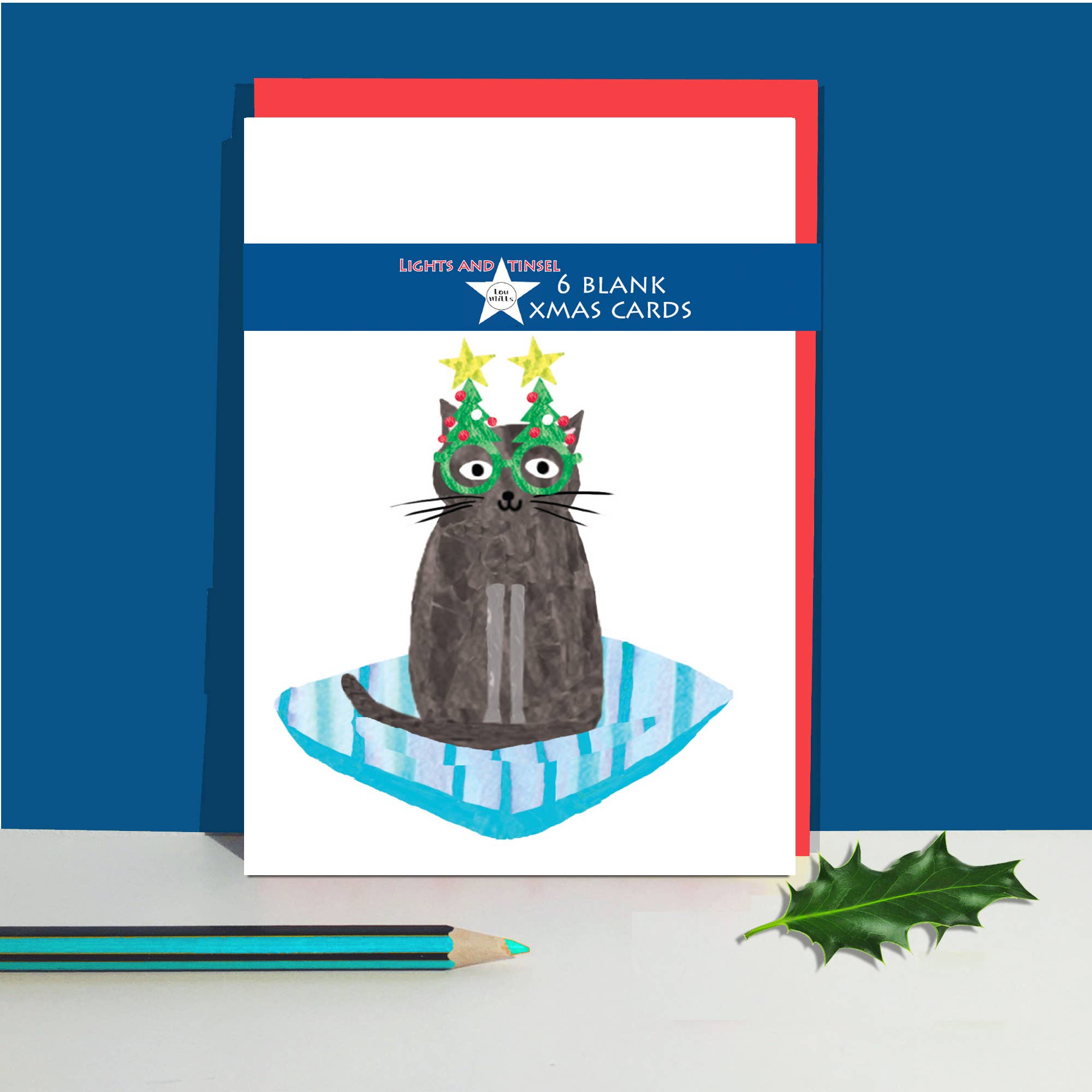 A pack of 6 blank Christmas cards, featuring a whimsical illustration of a dark grey cat wearing a green Christmas tree hat and a blue striped blanket, displayed on a white surface with a teal pencil and a holly leaf against a dark blue background.