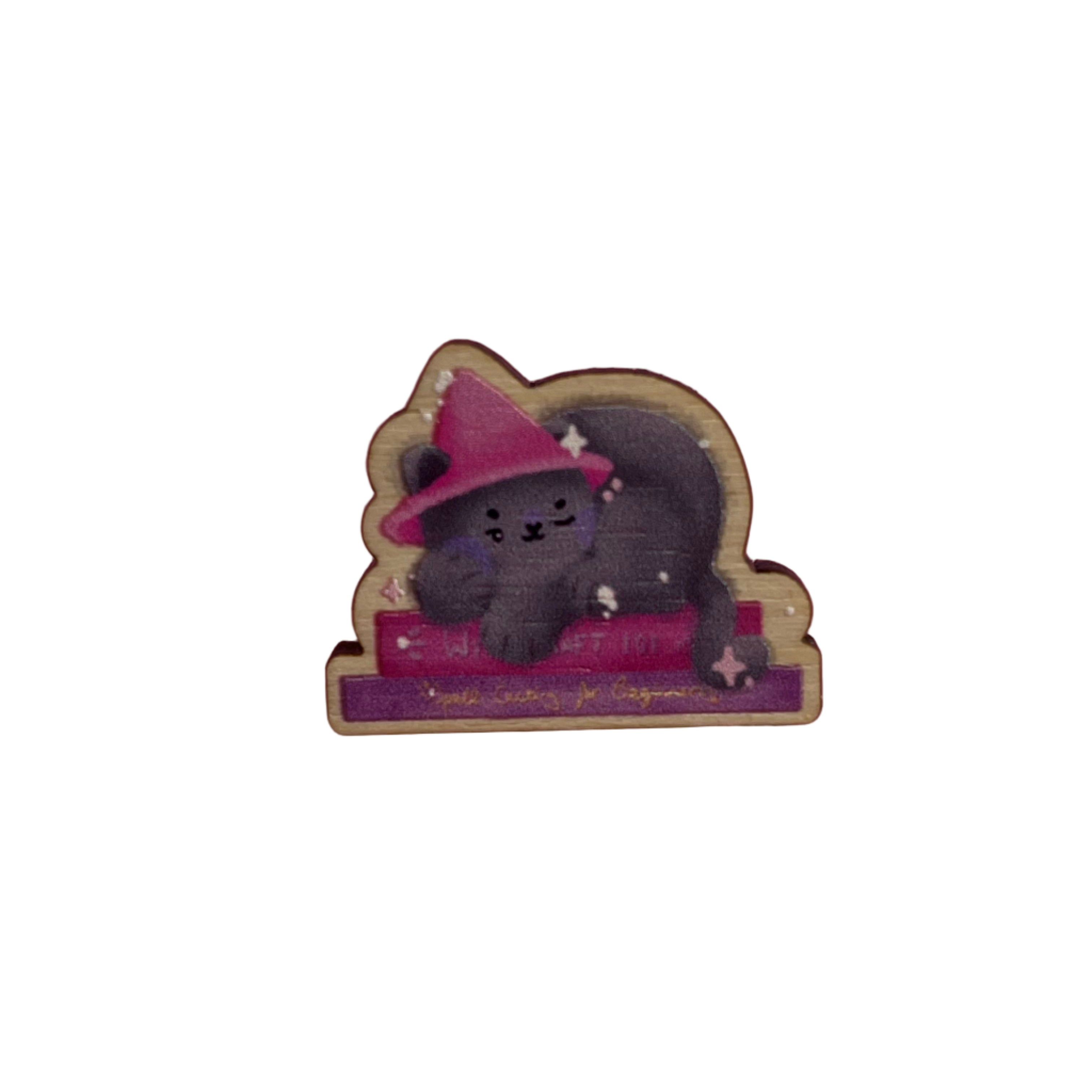 Wooden pin badge of a grey cat wearing a pink witch's hat, sleeping on a stack of books. Book titles are 'Witchcraft 101' and 'Spellcasting for Beginners'. This badge is printed onto 3mm thick responsibly sourced European birch plywood.