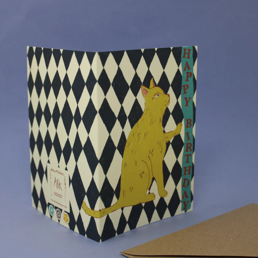 Birthday card with a cat illustration on a black and white checkered background, sat on a purple background with a brown envelope.