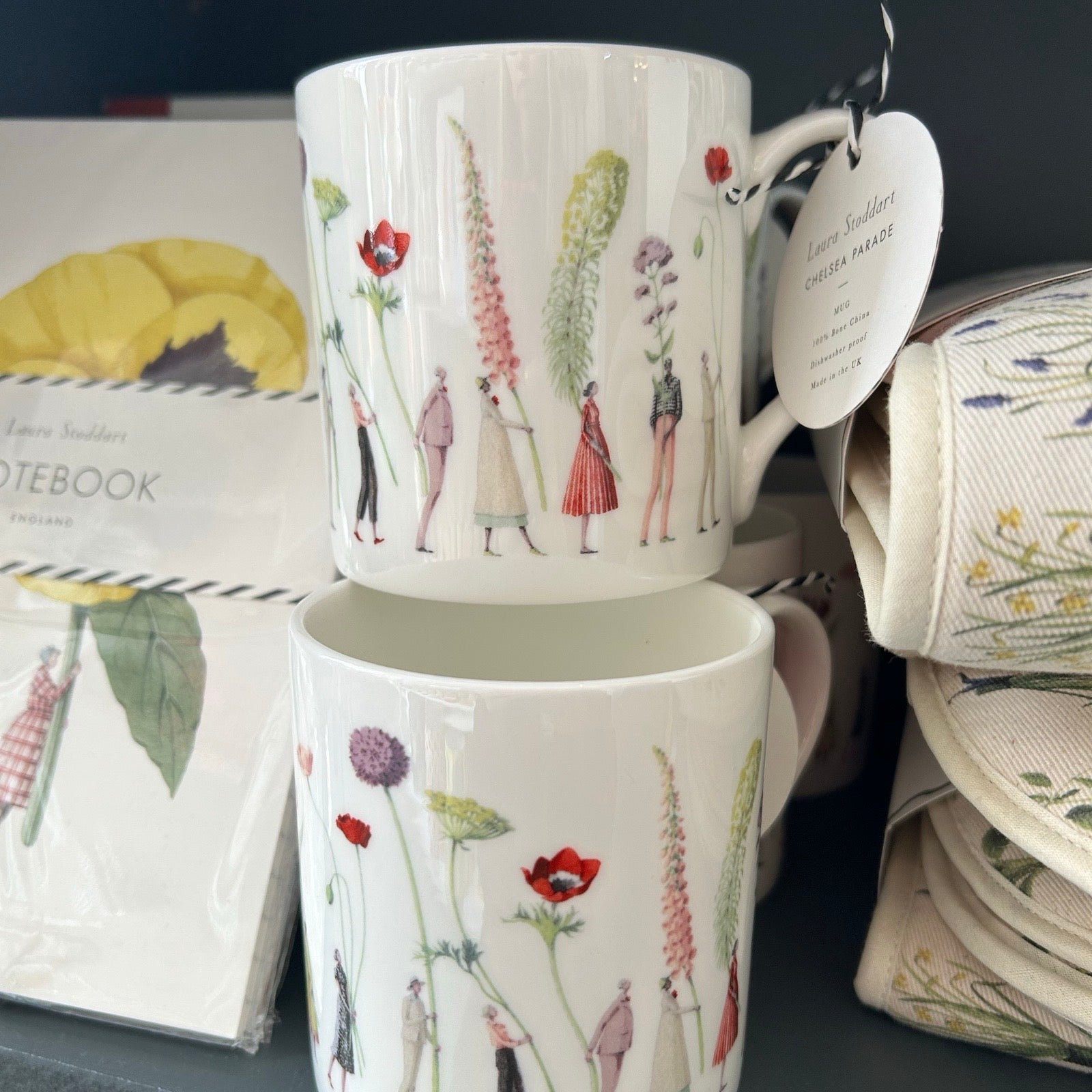 Two ceramic mugs with floral and figure design on a shelf with stationery items.