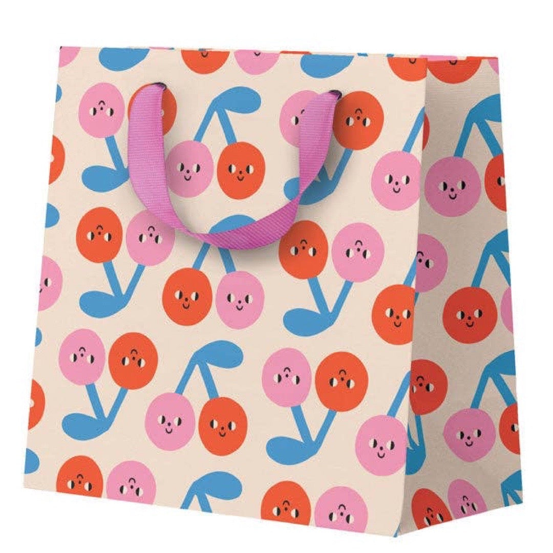 A paper gift bag with a repeating pattern of smiling cherries in pink, orange, and blue on a cream background, featuring purple ribbon handles.