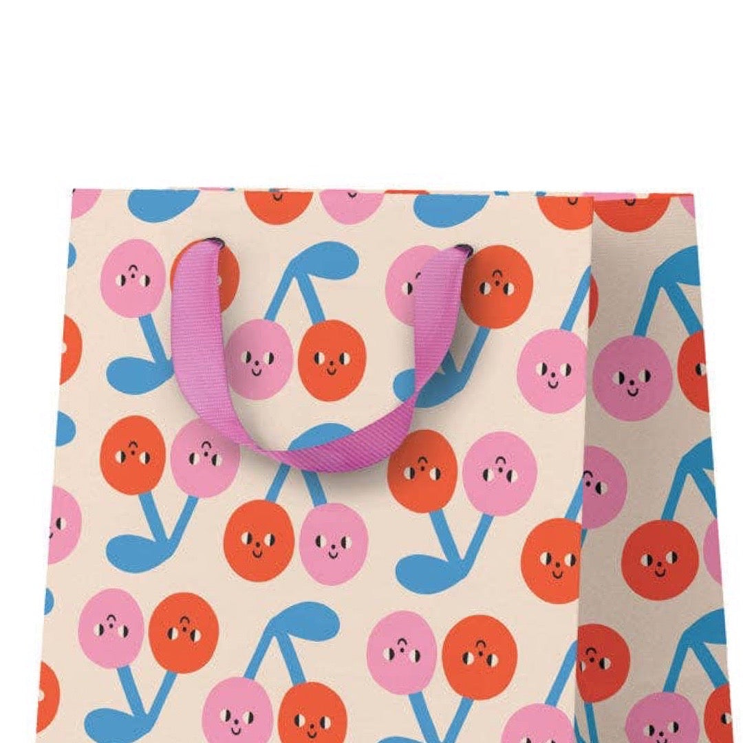 A gift bag with a repeating pattern of smiling cherries in pink, orange, and blue on a cream background, featuring purple handles.