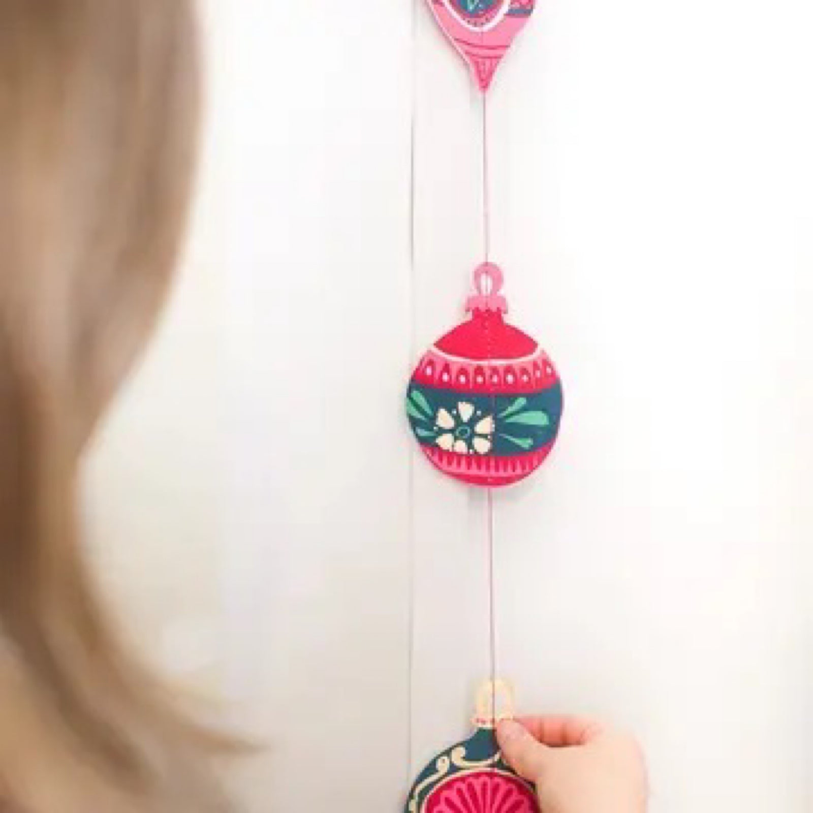 A vertical garland with red, pink and green Christmas baubles hanging against a white wall.