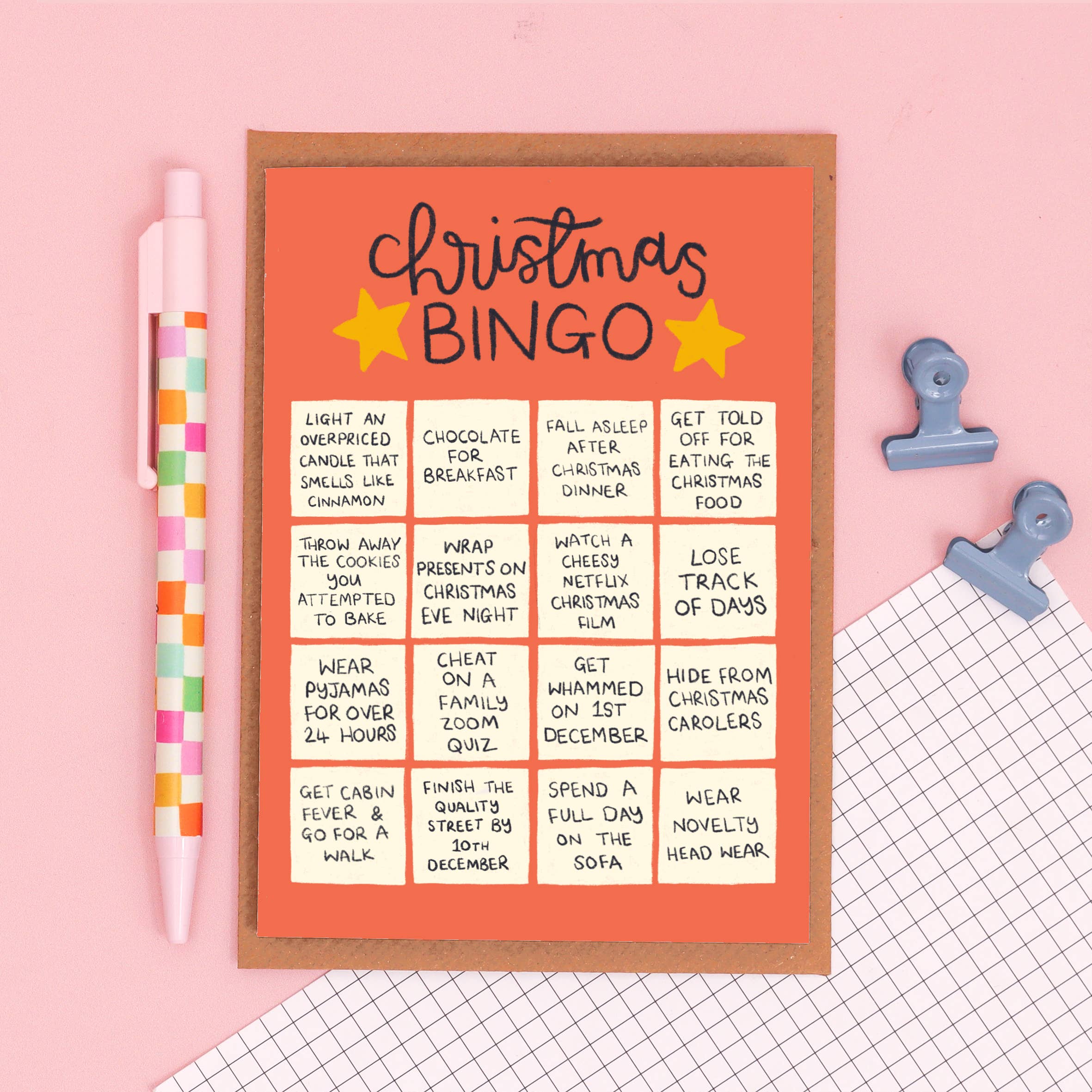 A Christmas Bingo game board with a warm orange background and black lettering, styled on a pink surface with a pink-and-white striped pen, a blue stationary, and a grid-patterned paper corner.