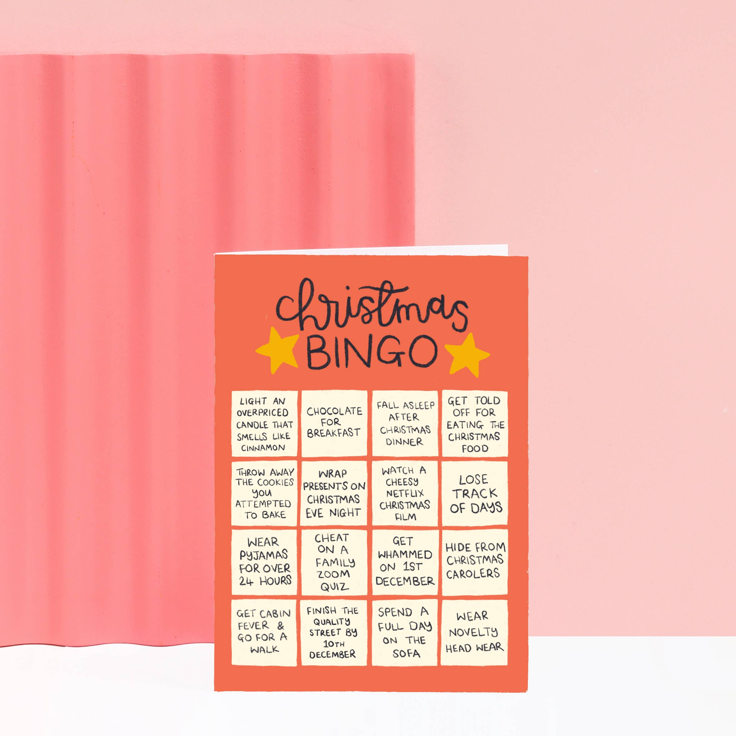 A Christmas Bingo card with a coral-orange background, black handwritten-style title, and yellow star accents, shown standing upright against a soft pink background.