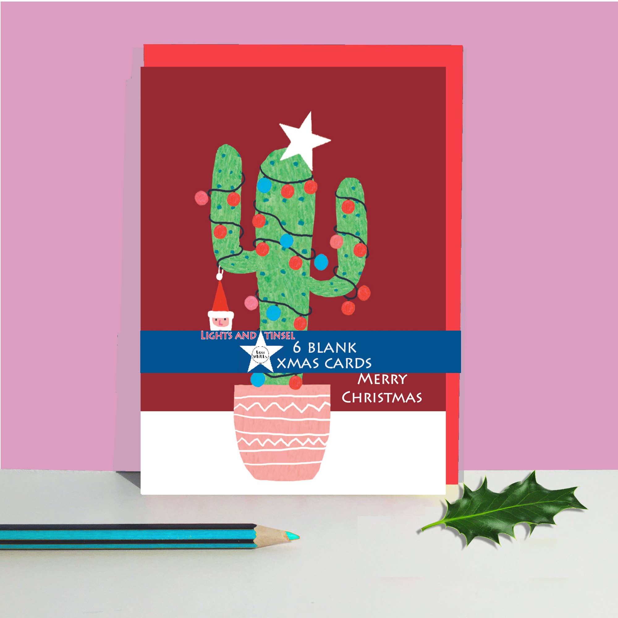 A Christmas card featuring a green cactus in a pink pot with red ornaments and a white star, shown standing upright on a white surface against a pink background. A blue pencil and a green holly leaf are placed in front.