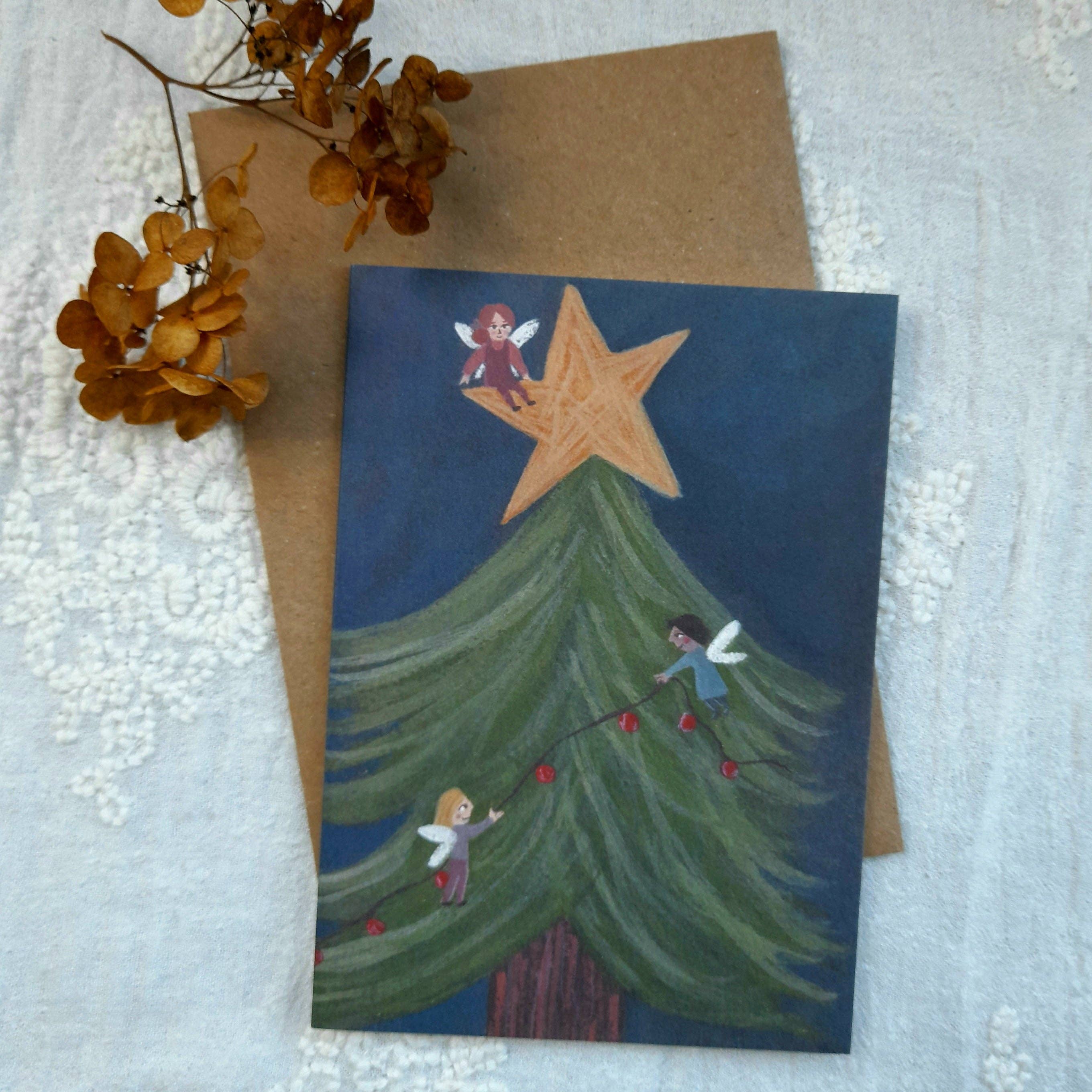 Christmas-themed greeting card with fairies decorating a tree, placed on a textured surface with a brown envelope.