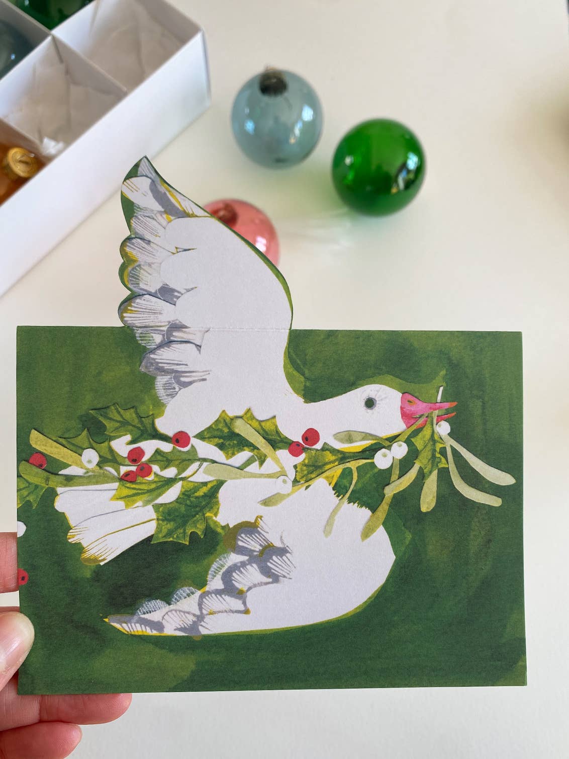 Card with a goose holding mistletoe and holly leaves, held by a hand on a white surface.
