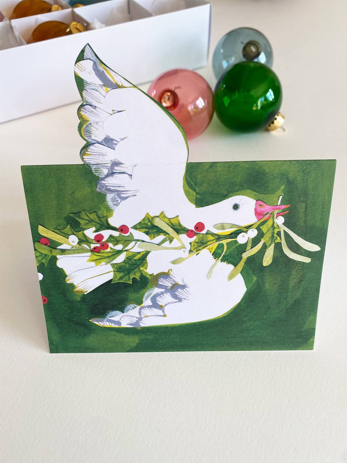Greeting card with a goose holding mistletoe on a green background, on a table next to baubles.
