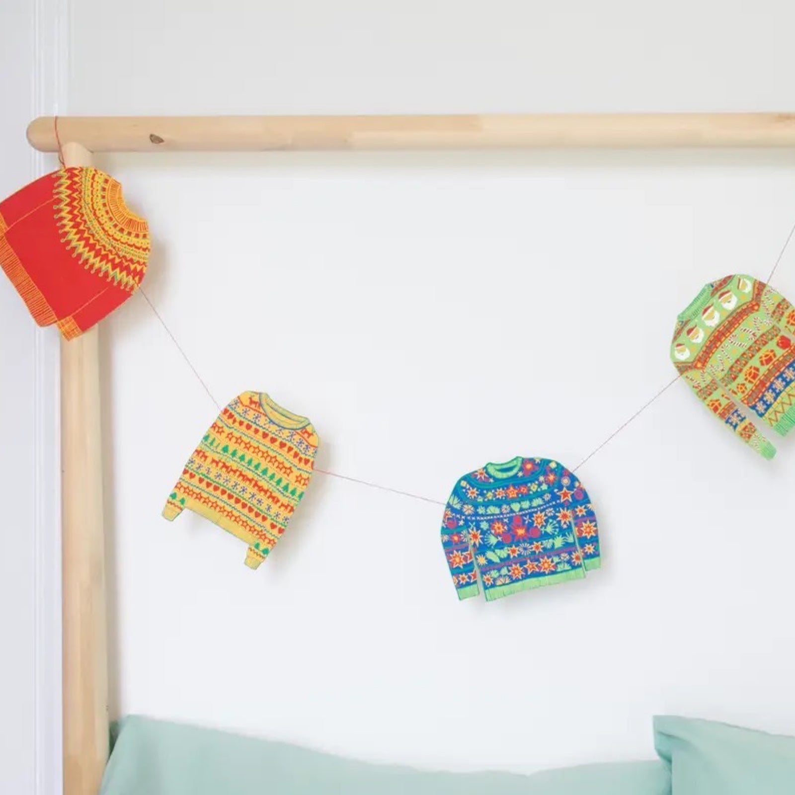 Christmas jumpers paper garland, featuring Christmas jumpers in red, green and blue patterns hanging from a wooden bed frame.