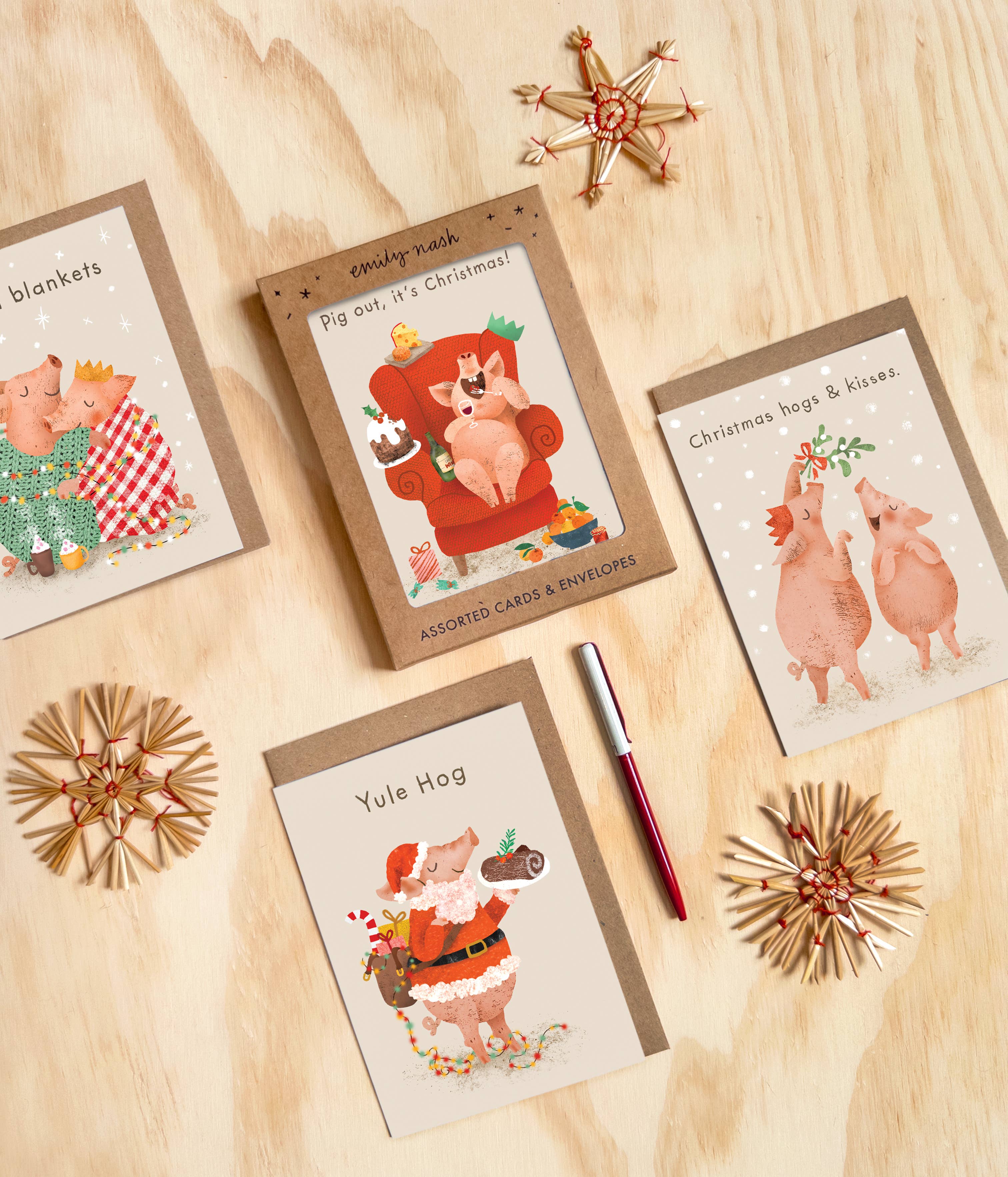 An assortment of Christmas-themed pig cards and blank cards arranged on a light wood surface with decorative star ornaments and a red pen. The cards feature illustrated pigs in holiday outfits with phrases like 'Pig out, it's Christmas!' and 'Yule Hog'.