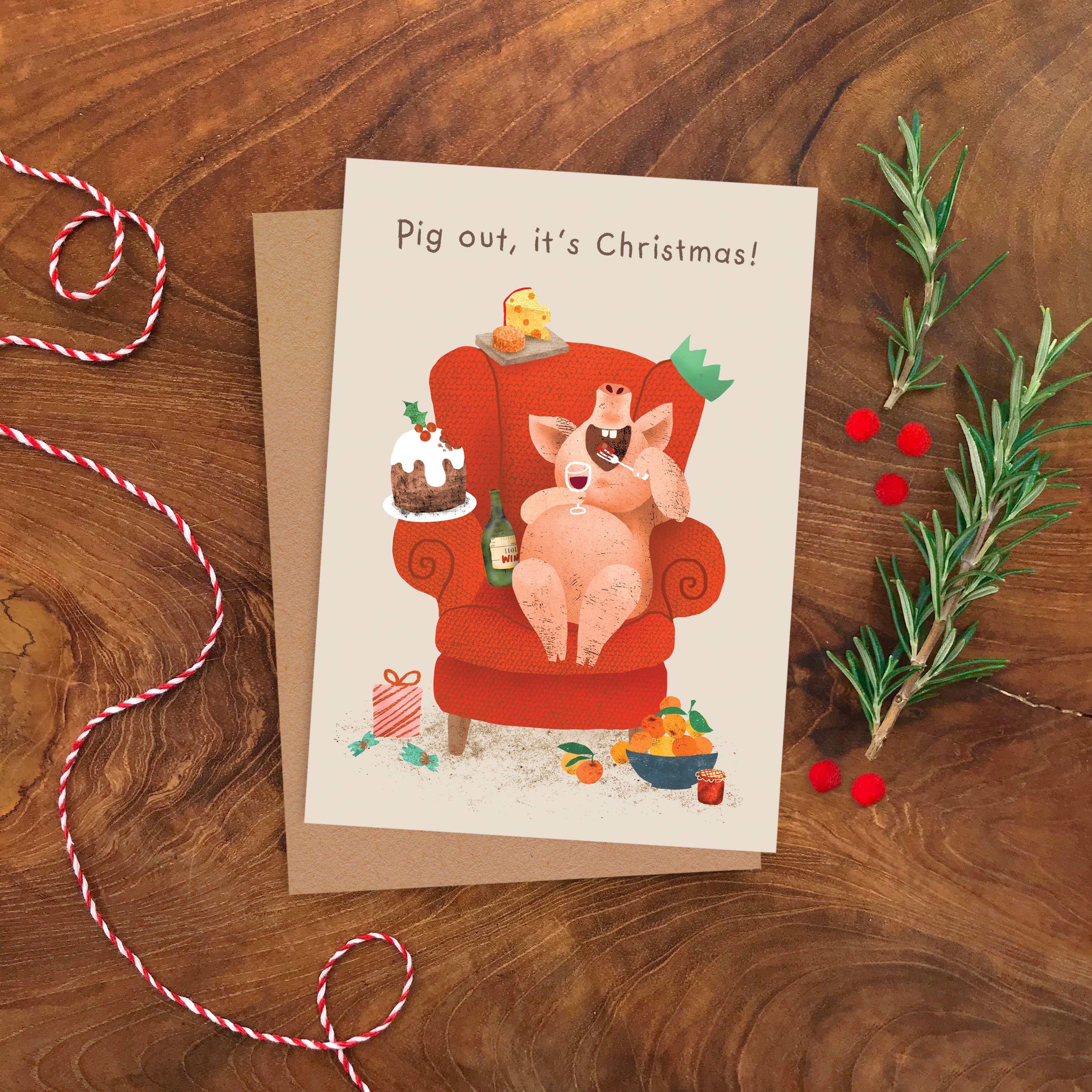 A Christmas greeting card featuring a cartoon pig relaxing in an orange armchair with the text 'Pig out, it's Christmas!' on a kraft envelope, styled on a wooden tabletop with bakers twine and rosemary sprigs.