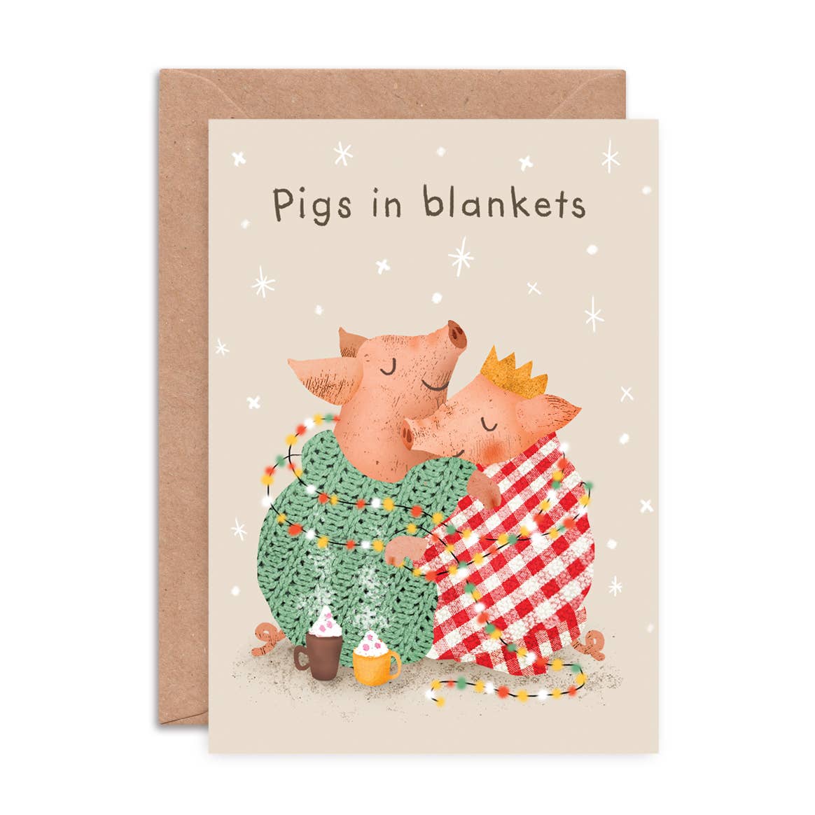 A greeting card with a kraft envelope, featuring the text 'Pigs in blankets' above an illustration of two pigs hugging under cozy patterned blankets.