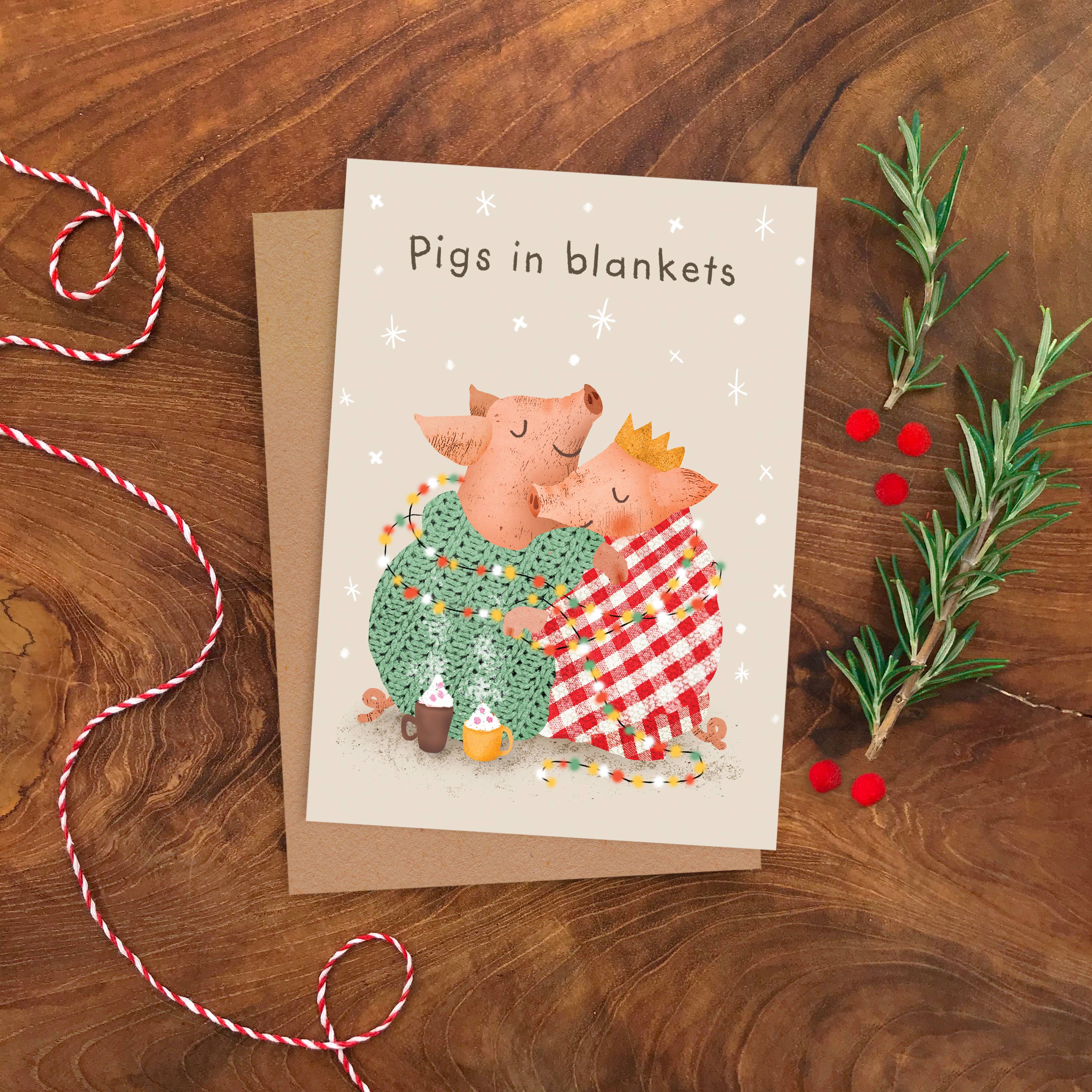 A greeting card with the text 'Pigs in blankets' and an illustration of two pigs hugging under string lights, styled on a wooden surface with a kraft envelope, red-and-white string, and rosemary sprigs.