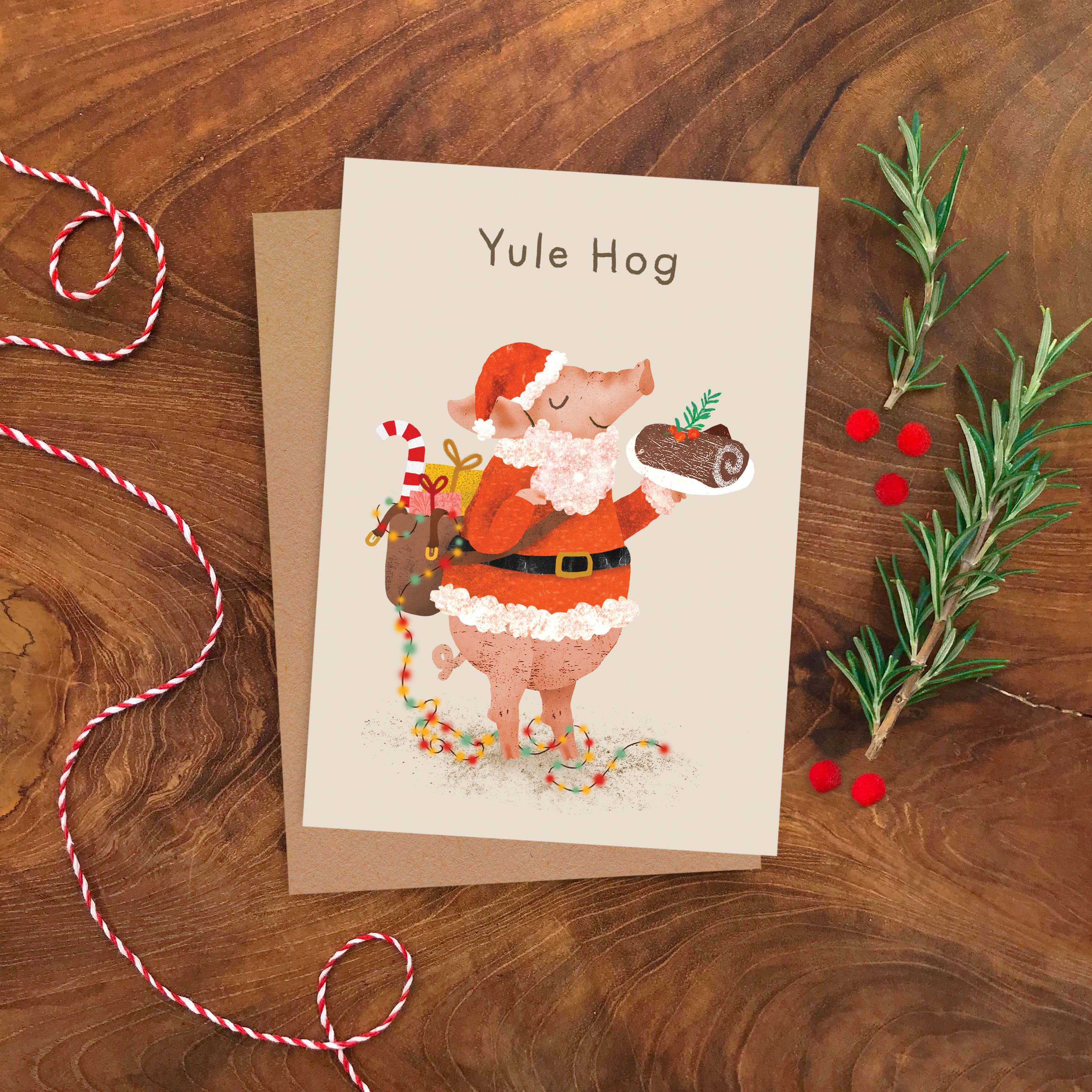 A greeting card featuring a whimsical illustration of a pig dressed as Santa Claus, labeled 'Yule Hog,' placed on a brown kraft envelope over a wooden surface with holiday props including red-and-white string and rosemary sprigs.