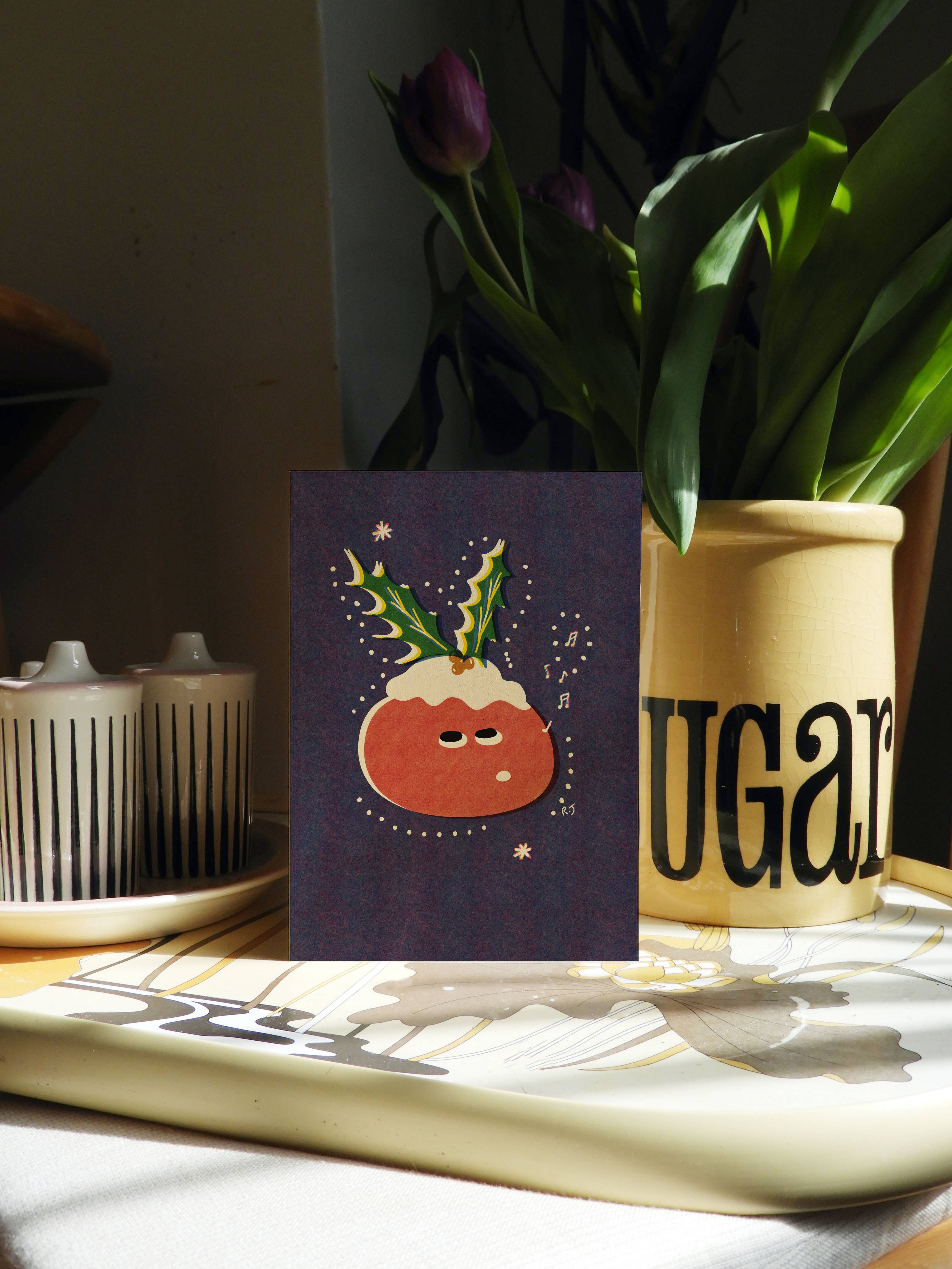 A greeting card featuring a whimsical illustration of a Christmas pudding with green holly leaves, set against a dark purple background. The card is styled on a tray next to a yellow ceramic vase with tulips and candle holders in a warm, sunlit interior setting.