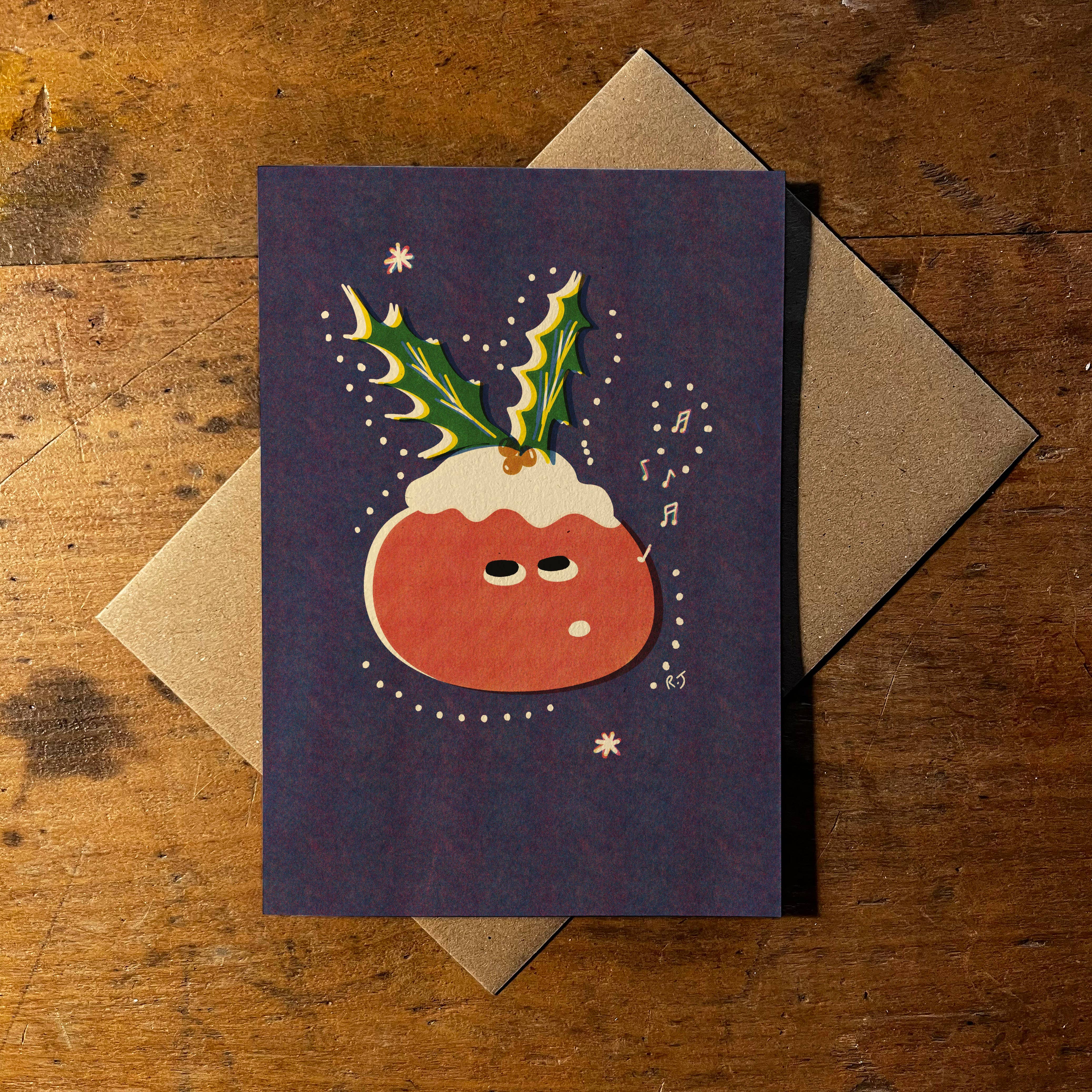 A Christmas greeting card featuring a cute, illustration of a pudding with holly leaves and snow, displayed on a rustic wooden table with a brown kraft envelope behind it.