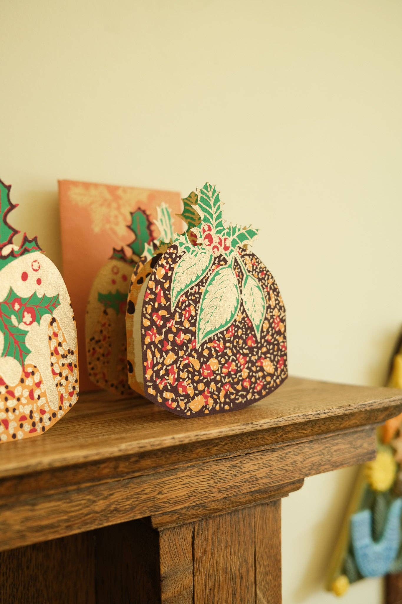 A decorative Christmas pudding card sits on a rustic wooden mantelpiece against a warm cream-coloured wall. The card features a dark brown background with a dense red, green, and beige floral pattern, topped with green leaves and red berries.
