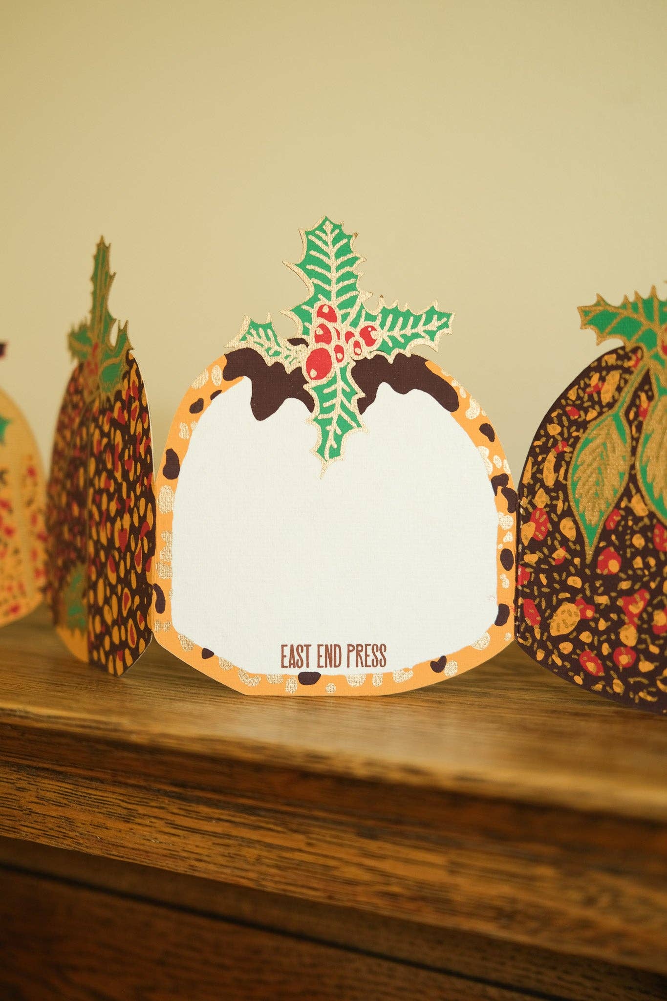 A close-up of a decorative card or sign on a wooden surface, with the words 'EAST END PRESS' printed near the bottom. The card has a white centre, an orange border, and a holly motif at the top, with other patterned cards partially visible in the background.