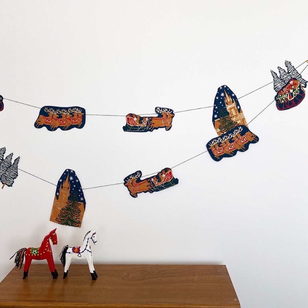 A skyline paper garland featuring Christmas trees, sleighs and reindeer hanging against a white wall.