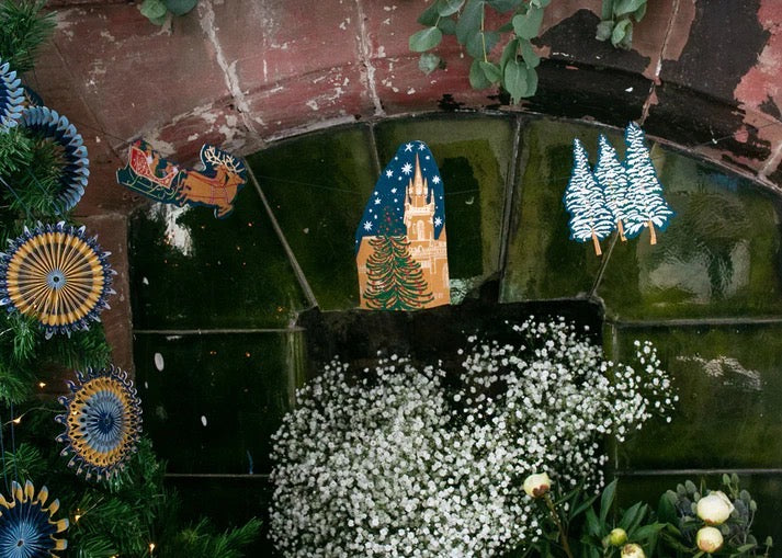 A skyline paper garland featuring Christmas trees, sleighs and reindeer hanging against a fireplace with green plants.