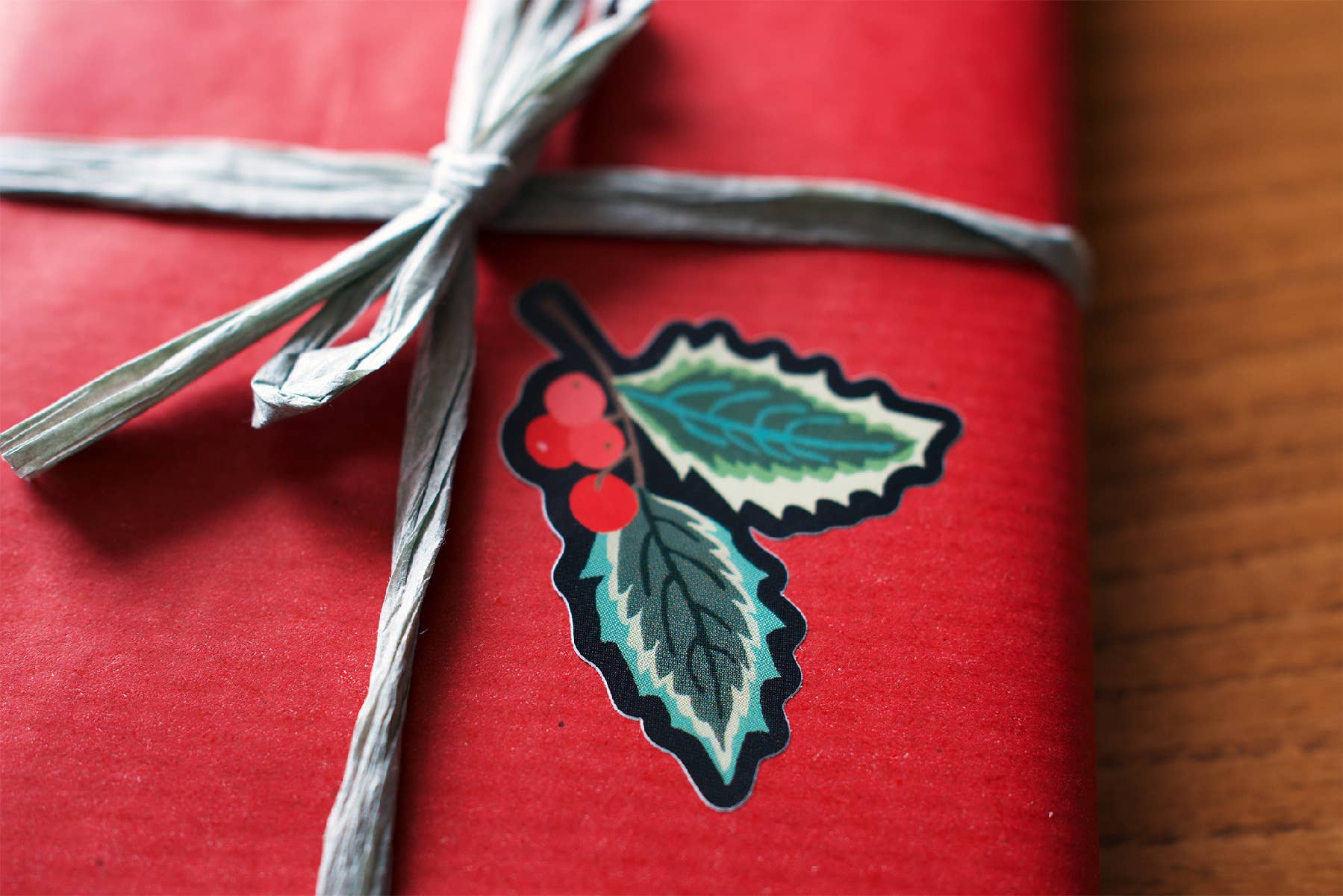 A close-up of a red wrapped gift tied with a grey ribbon, featuring a die-cut sticker of holly leaves and red berries on the front.