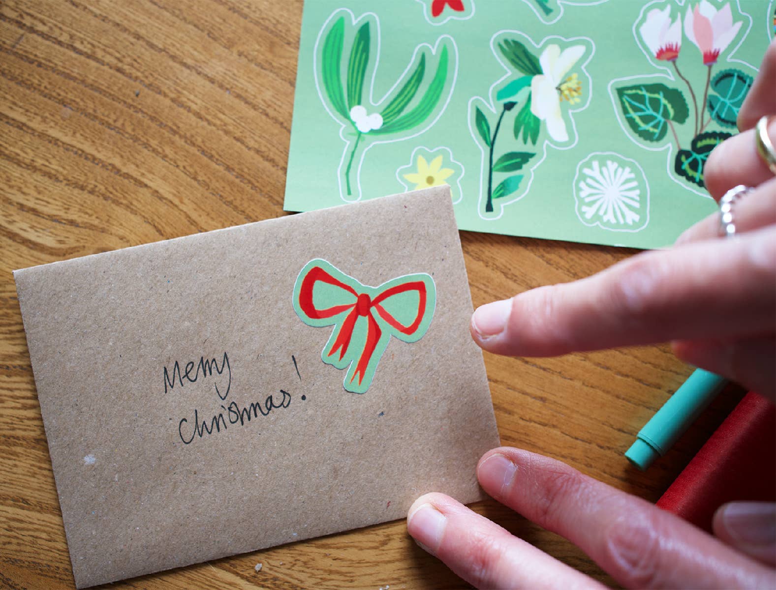 A close-up of a hand holding a brown kraft paper Christmas card with 'Merry Christmas!' written on it and a red bow illustration. A sheet of floral stickers is visible in the background on a wooden table.
