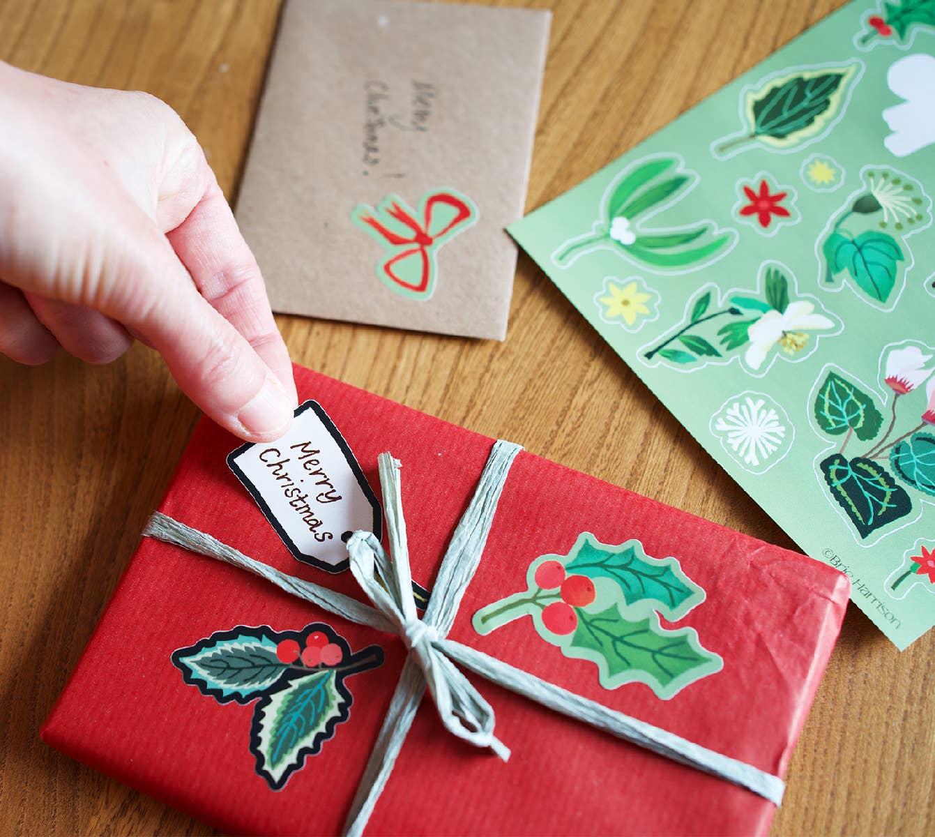 A hand is wrapping a gift box with red paper and a twine bow, decorated with holly leaf stickers. A kraft paper card and a sheet of botanical stickers are visible on a wooden table.