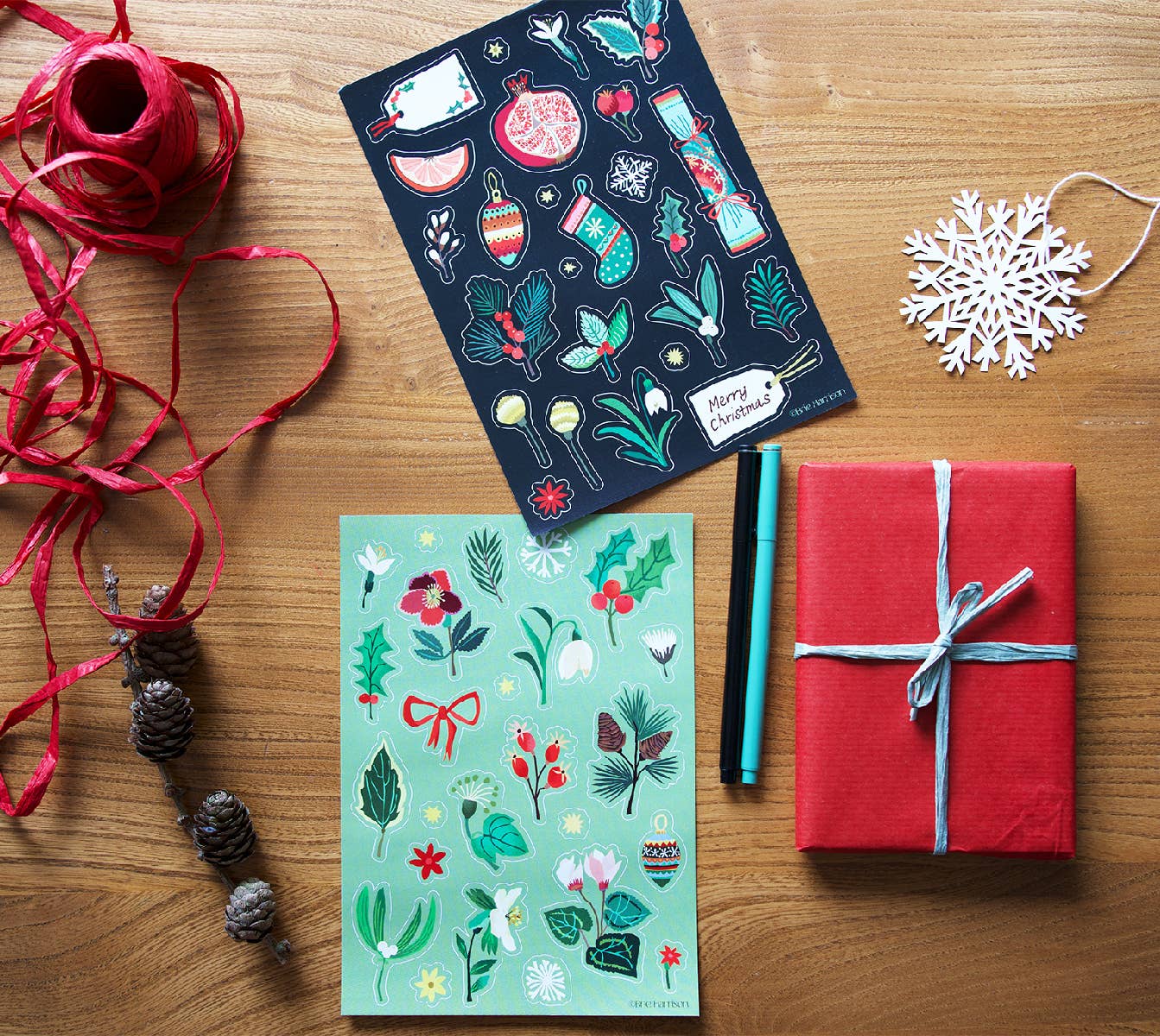 Two decorative holiday sticker sheets displayed on a wooden tabletop alongside festive props, including red wrapping ribbon, pine cones, a white snowflake ornament, and a wrapped red gift box with a blue ribbon.