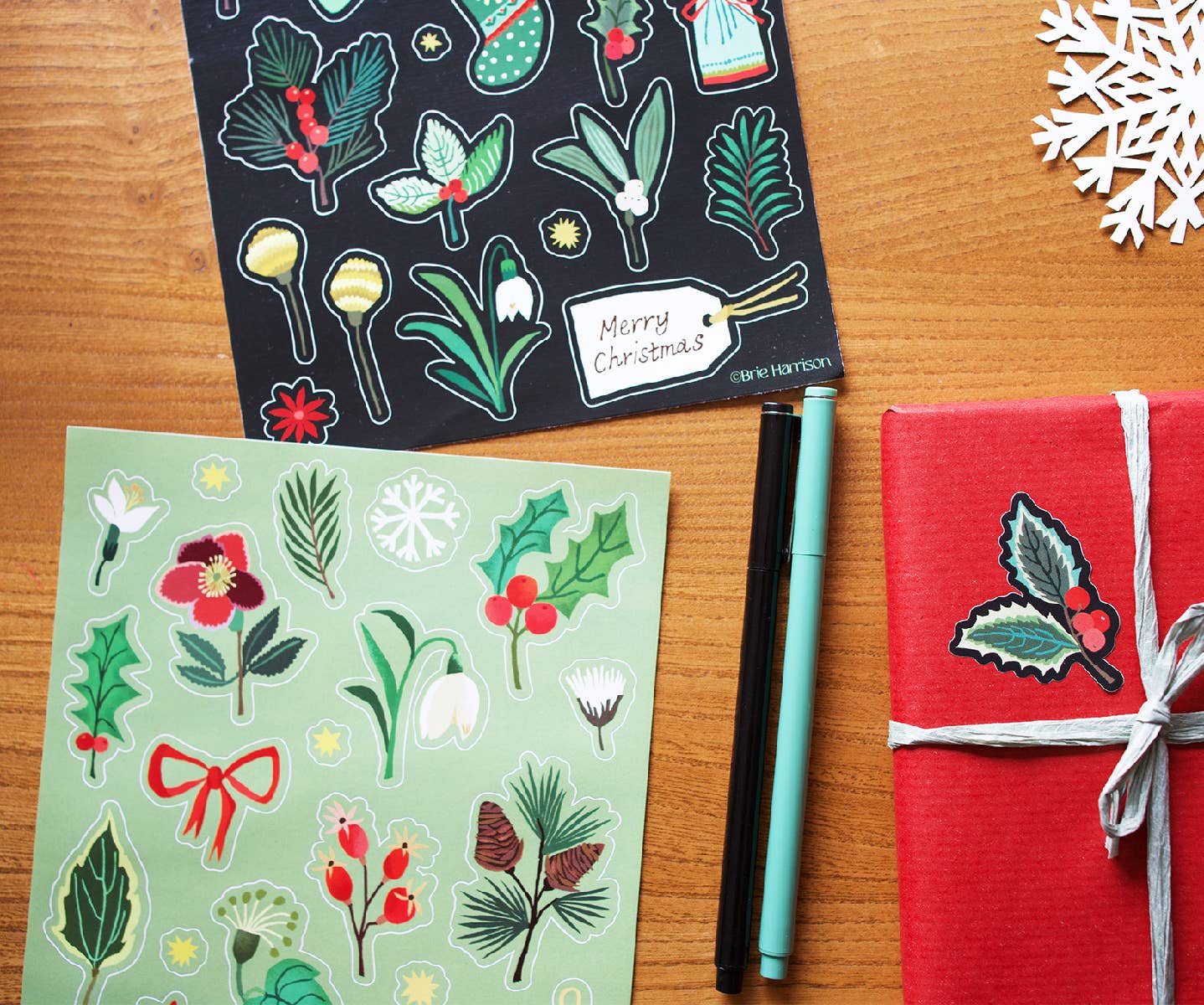 Two Christmas-themed sticker sheets on a wooden tabletop, with one black sheet featuring winter botanical illustrations and a mint green sheet with holly, berries, snowflakes, and bows. A wrapped red gift box with a holly sticker and gray ribbon sits to the right, alongside two pens and a white paper snowflake.