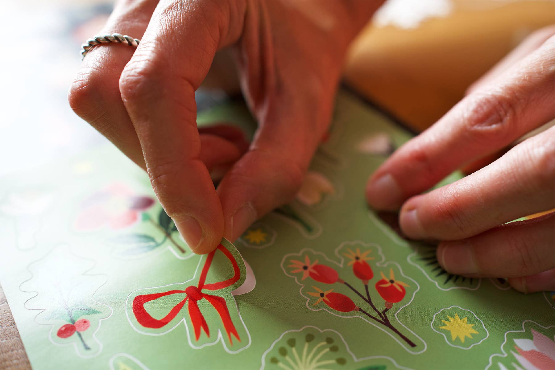 Close-up of a person's hands peeling a red bow-shaped sticker from a sheet of colourful stickers featuring floral and botanical designs.