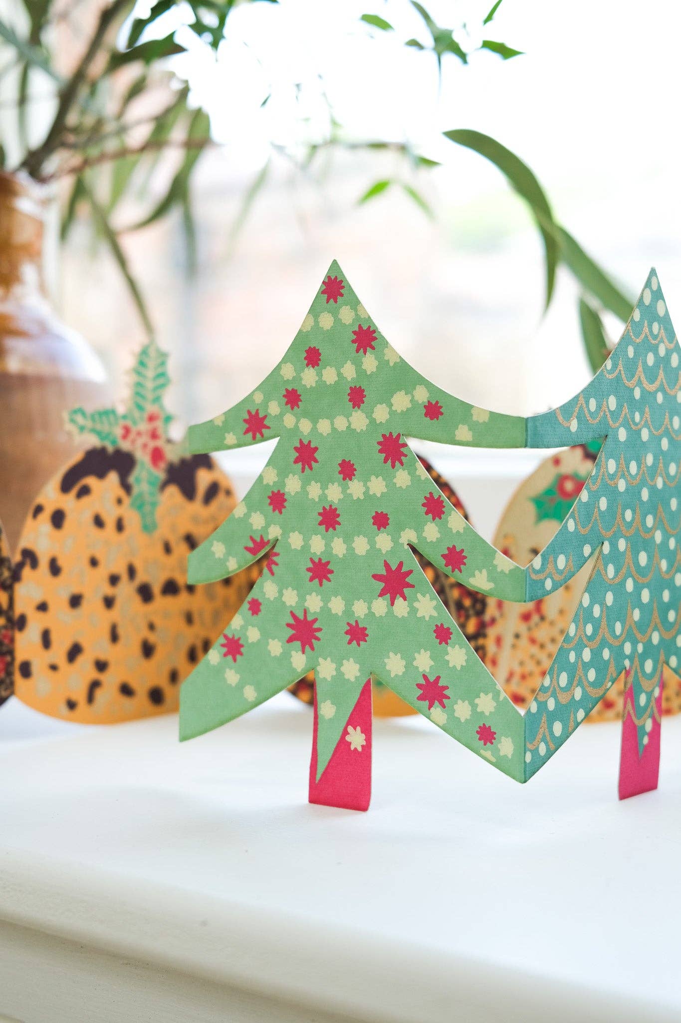 A decorative paper Christmas tree card in green with pink and cream star patterns, displayed on a white surface with a blurred background.