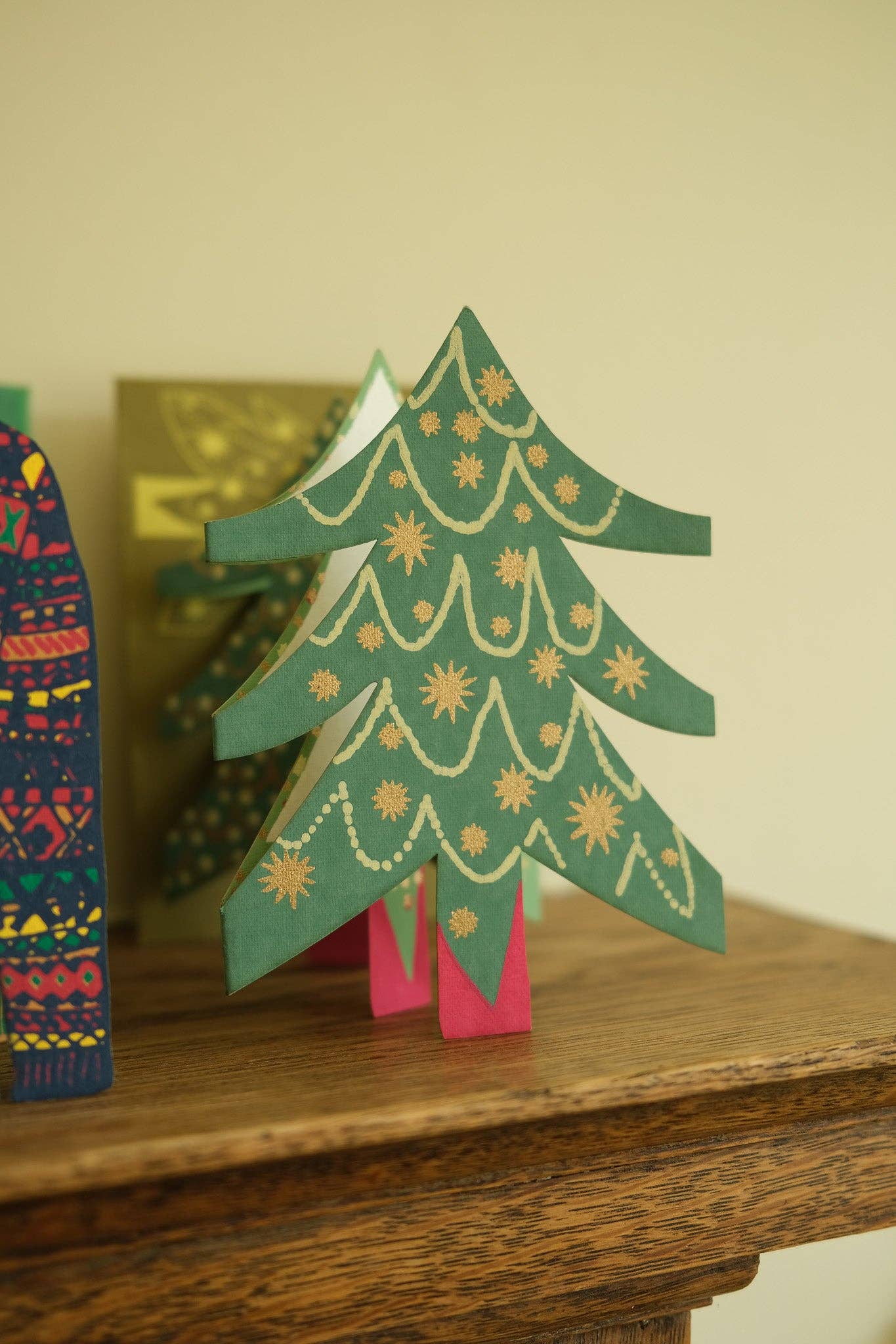 A green paper Christmas tree decoration with gold star and wave patterns, standing on a pink base on a wooden table.