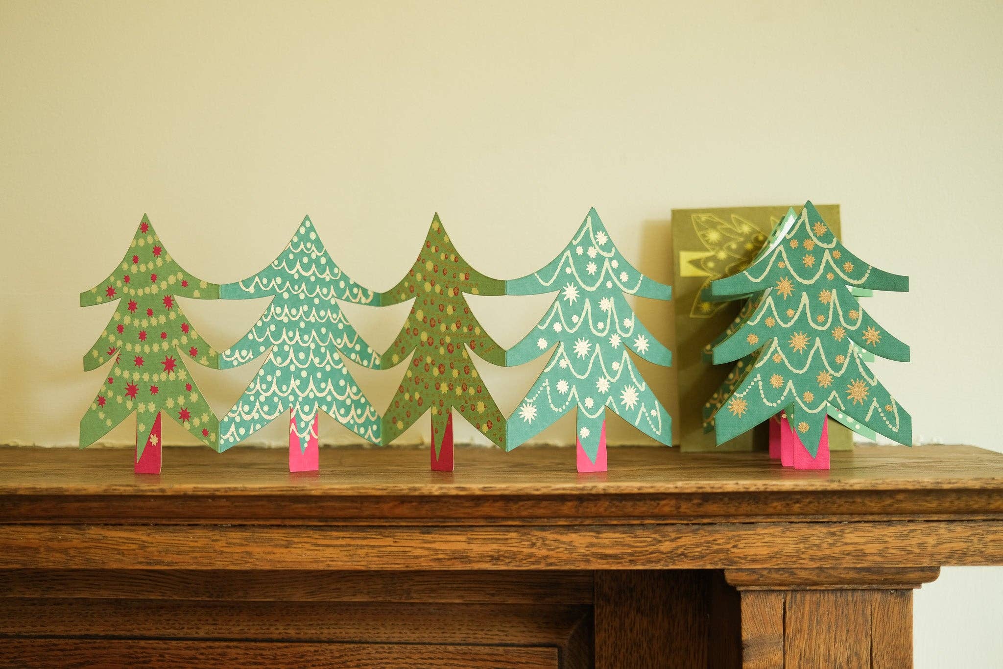 A set of five decorative paper Christmas tree ornaments in various green shades, decorated with white patterns and small red and gold accents, displayed on a wooden mantelpiece against a beige wall.