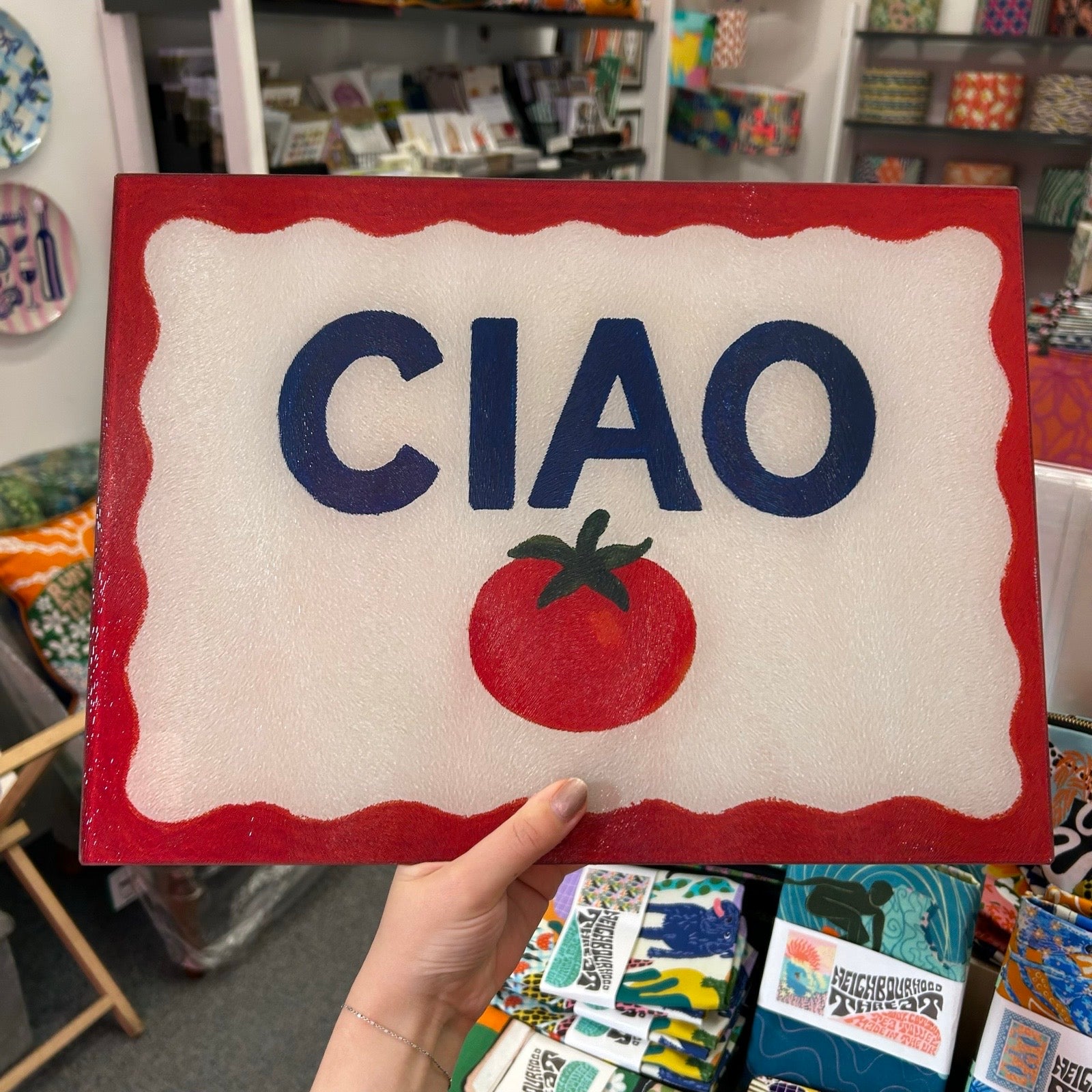 A hand holds a decorative chopping board with a red border, a white centre, the word 'CIAO' in large navy letters, and a red tomato graphic below. The background shows a blurred retail store interior with shelves and colourful items.