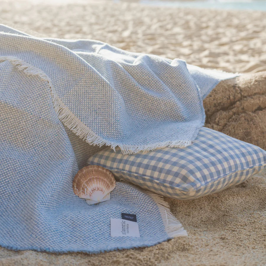 Light blue blanket and checkered pillow on sandy beach with shell.