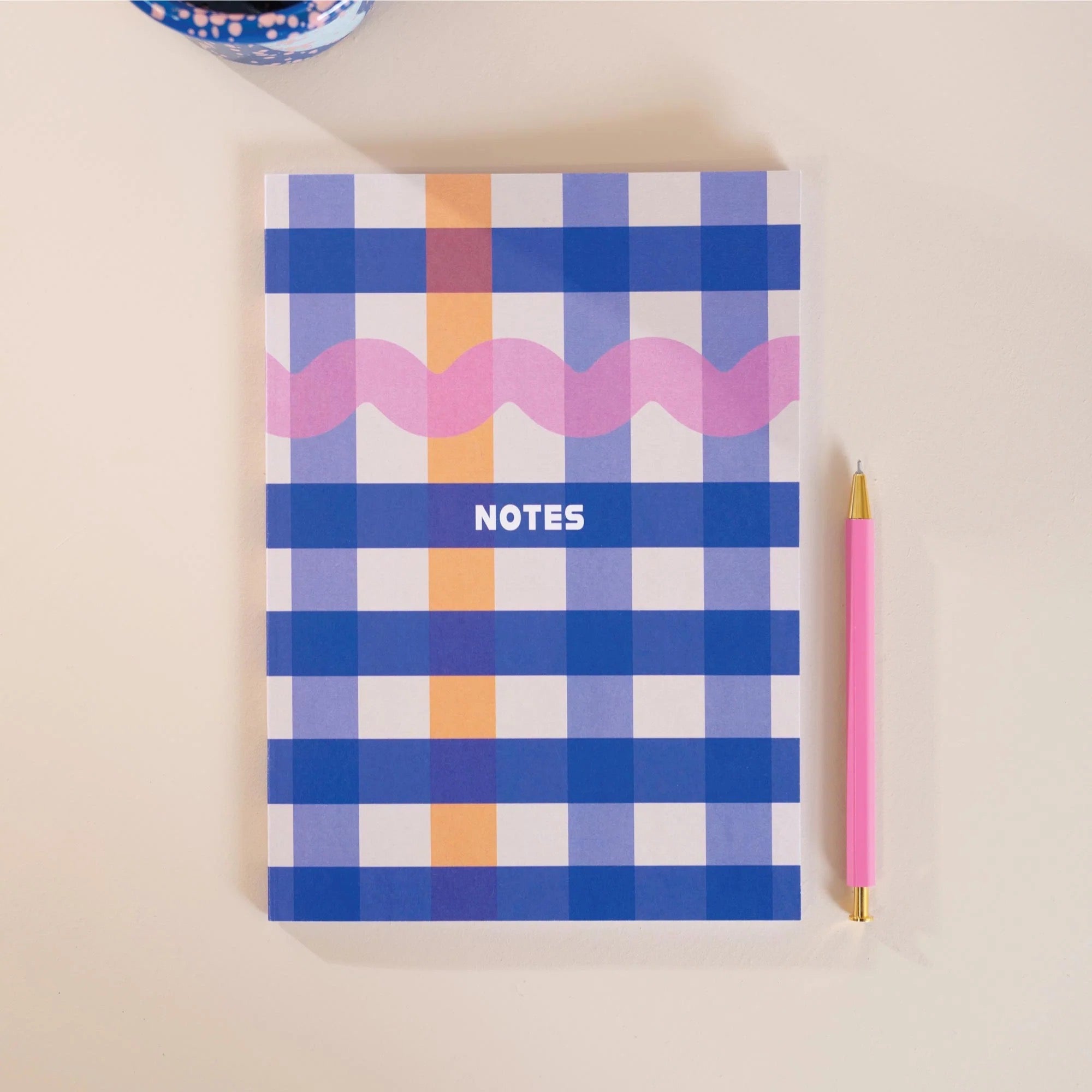 Notebook with a checkered design and wavy line, accompanied by a pink pencil on a beige background.