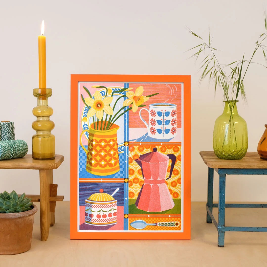 Framed artwork with colourful design of a pitcher, flowers, and kitchen items on a beige wall, next to plants and candles.
