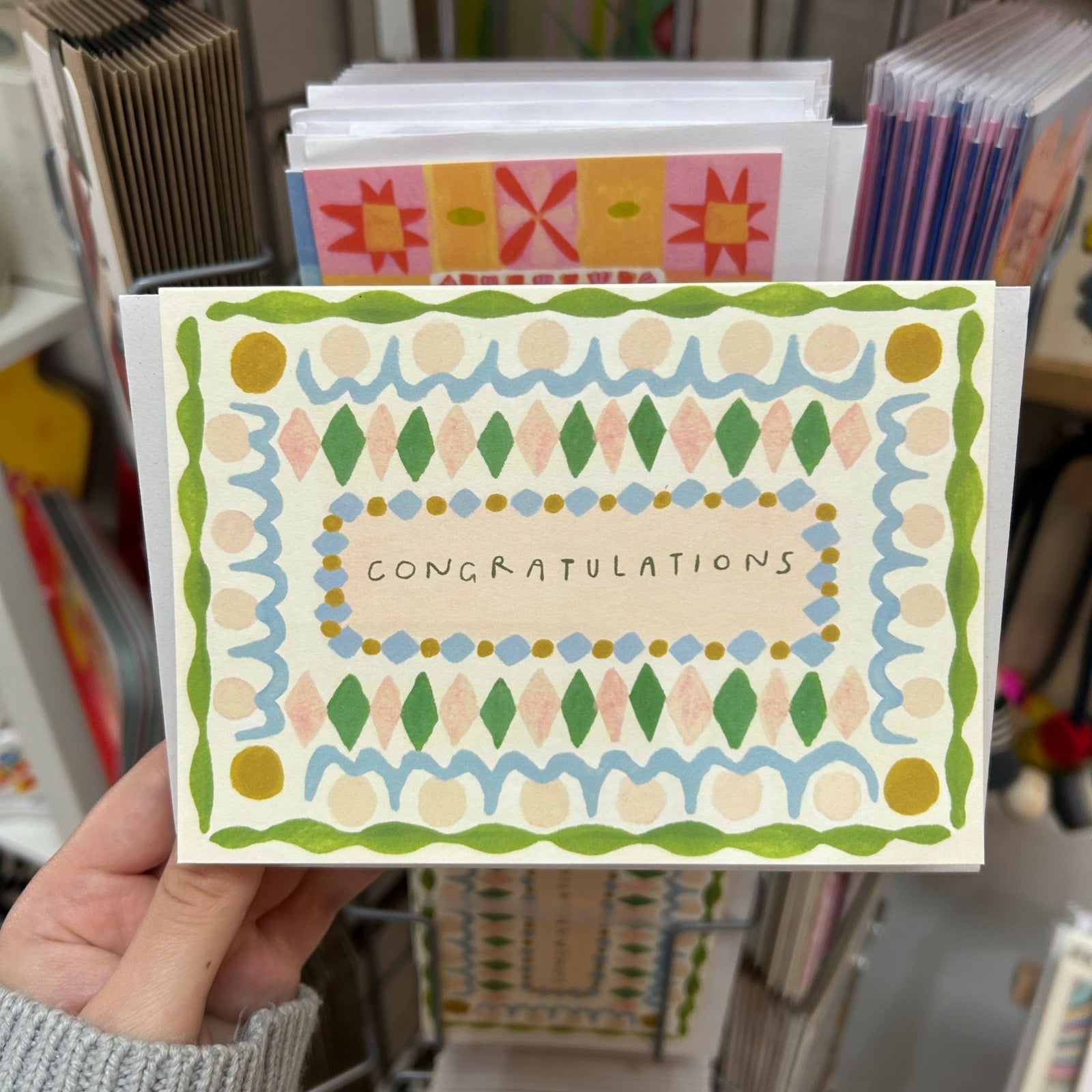 Hand holding a 'Congratulations' card with decorative patterns in a store setting.